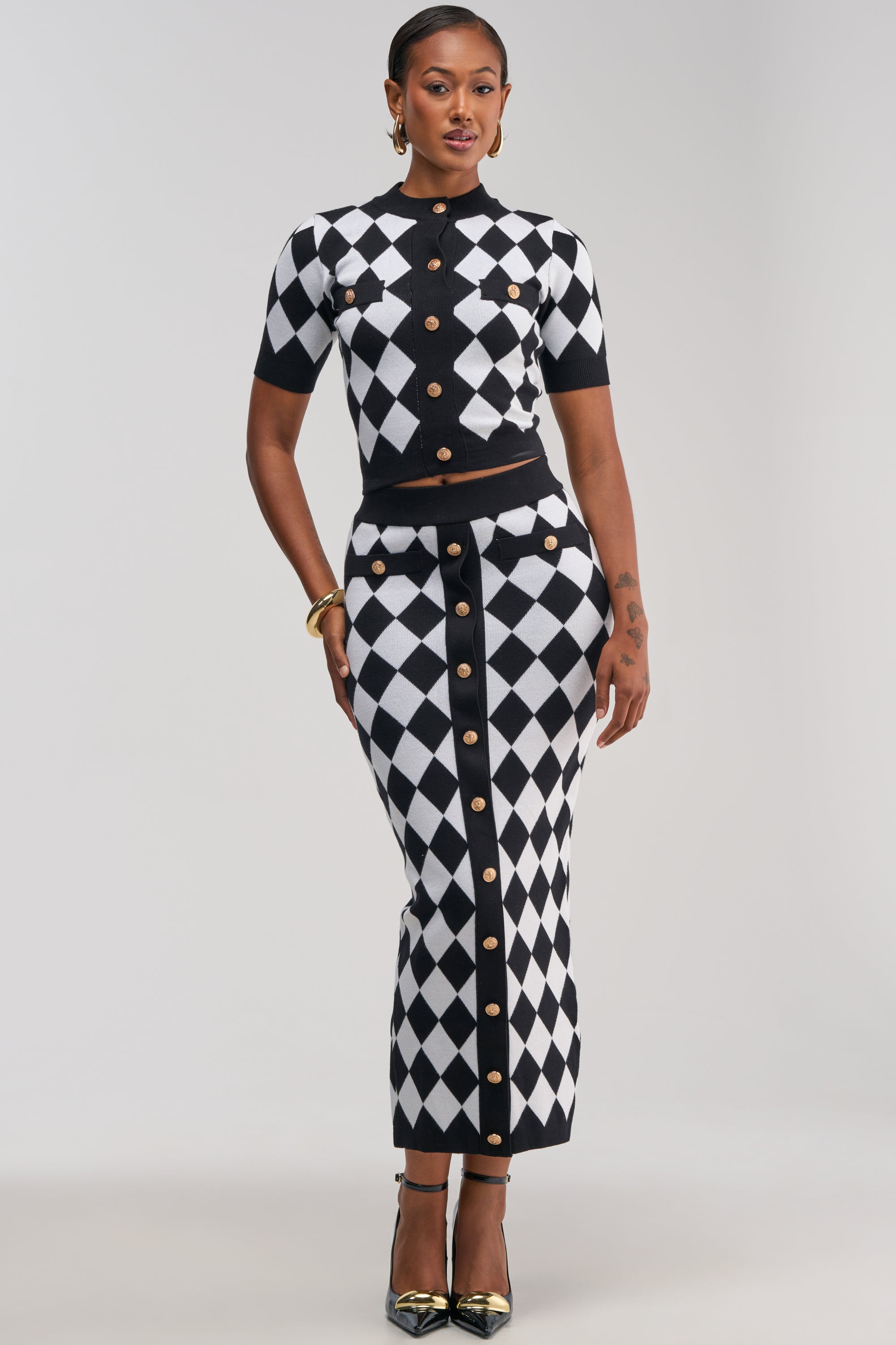A woman stands against a plain background wearing “THE BEST PART MAXI SKIRT,” a matching black and white argyle knit maxi skirt with gold buttons, paired with a coordinating short-sleeve top. She completes the look with hoop earrings, bangles, and pointed black heels.