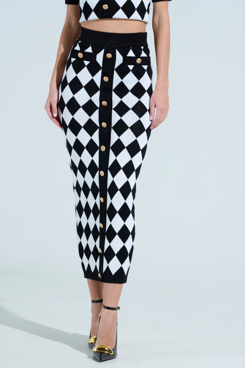 Someone wears THE BEST PART MAXI SKIRT, featuring a black and white checkered design with gold buttons in front. It's paired with a matching top and gold-tipped black heels, all against a light blue background.