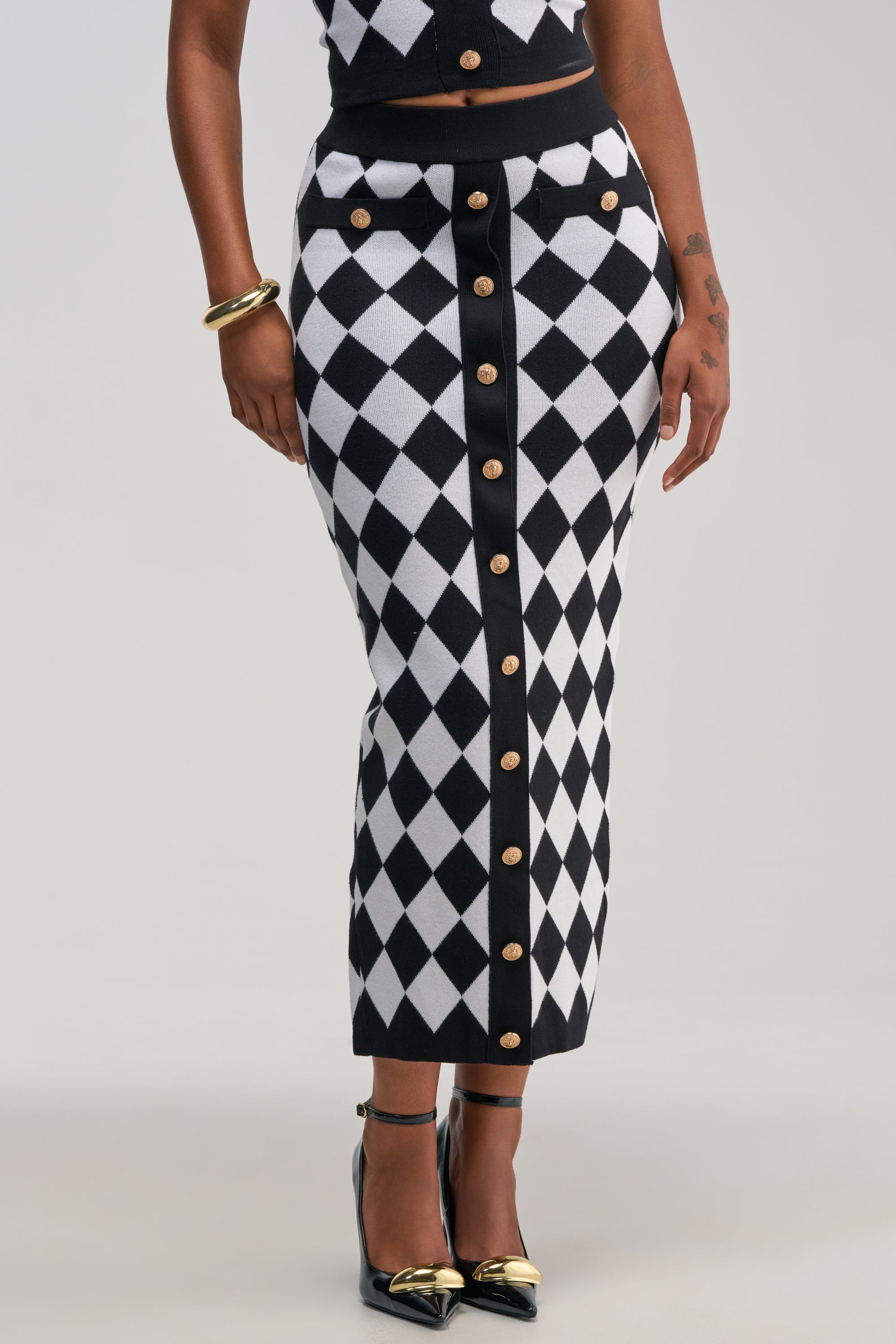 A person styled in THE BEST PART MAXI SKIRT—a high-waisted, black and white diamond-patterned maxi skirt with gold buttons down the front—paired with black heels featuring gold tips, a gold bangle, and showcasing tattoos on one arm.