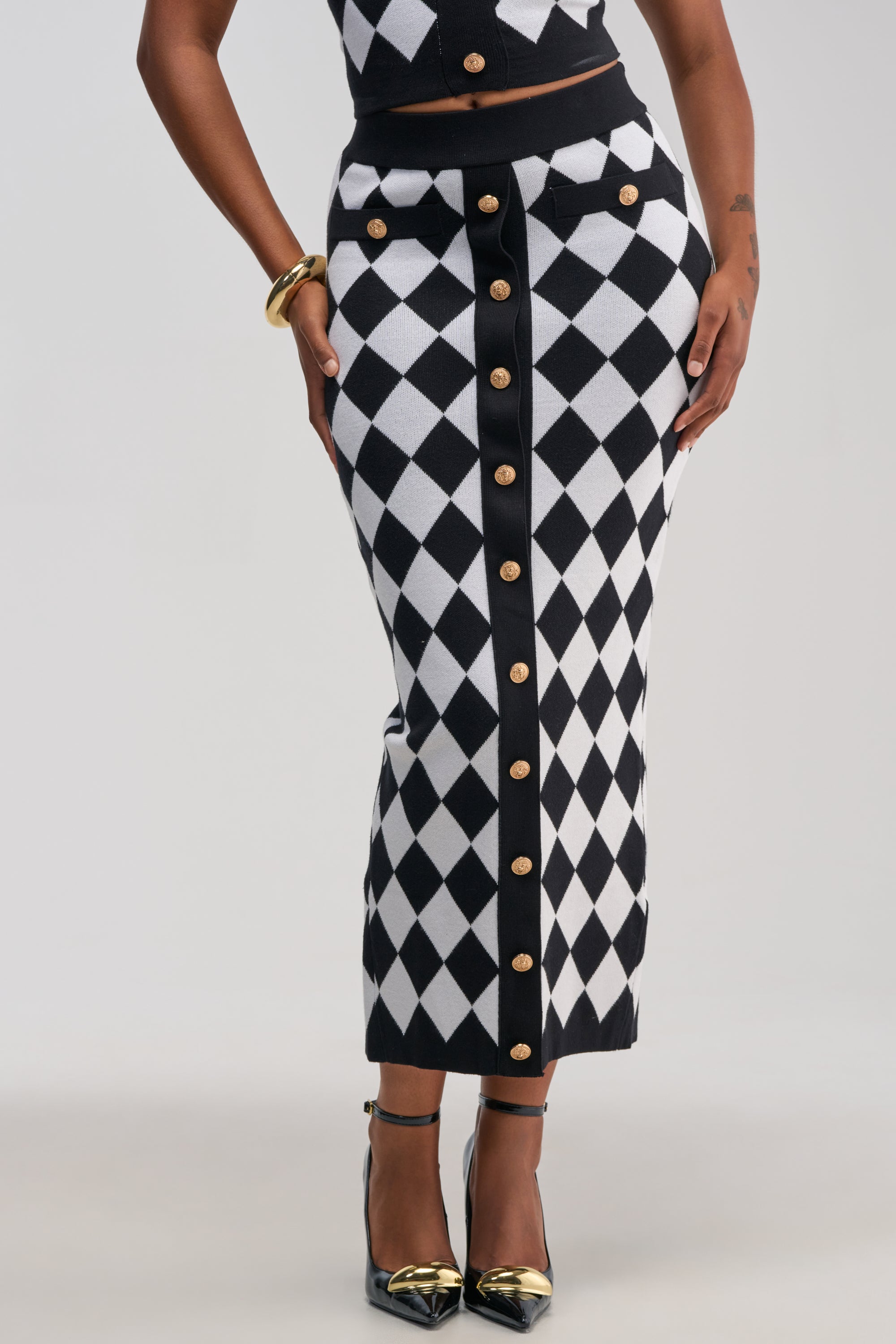 Wearing THE BEST PART MAXI SKIRT—a black-and-white diamond-patterned skirt with gold buttons down the front styled in a classic business casual look—she pairs it with black heels and gold bracelets.