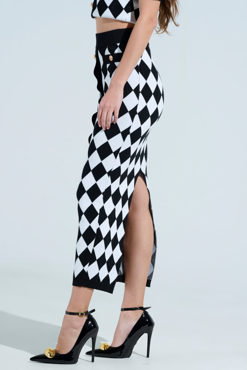 Side view of a person wearing "THE BEST PART MAXI SKIRT," showcasing a black and white checkered design with a side slit and matching crop top. The bodycon ensemble is paired with black high heels featuring gold details, set against a plain, light-colored background.