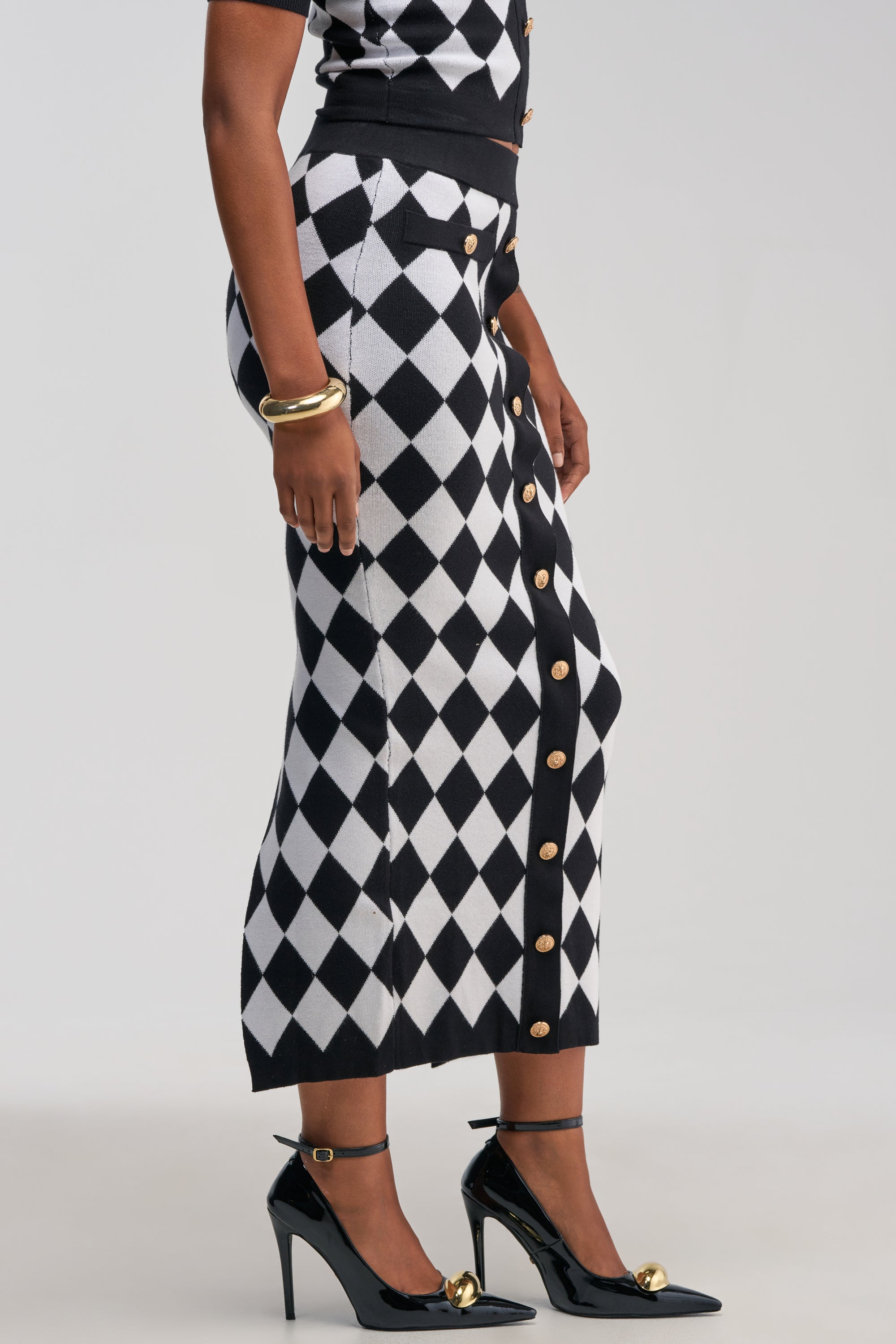 A person wearing THE BEST PART MAXI SKIRT—a black and white, diamond-patterned, button-front design styled in a business casual manner—paired with black high heels featuring gold accents and elegant gold bangles, stands against a plain background.