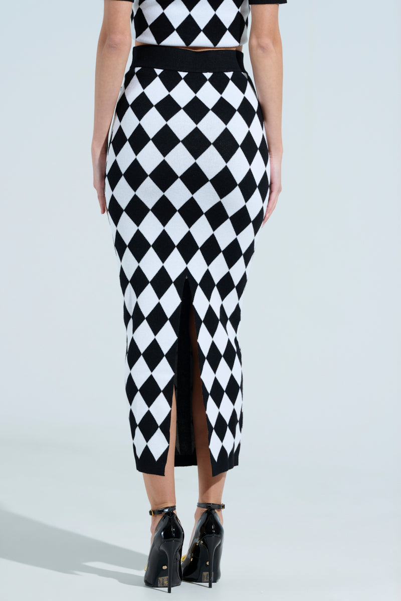 Someone is wearing AKIRA Label's THE BEST PART MAXI SKIRT, a high-waisted black and white checkered bodycon skirt with a back slit that extends below the knees. Paired with black ankle strap shoes, they stand against a light background.