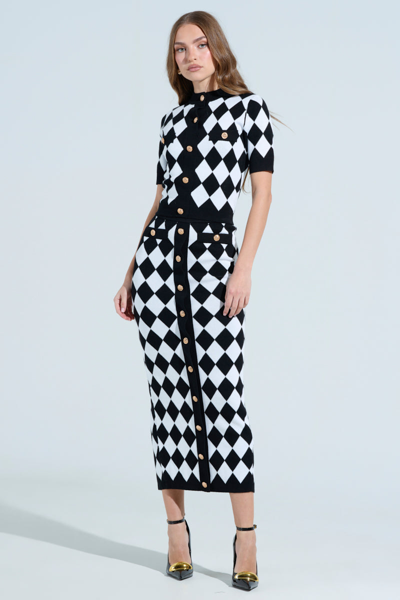 A woman in a chic black and white argyle outfit, featuring THE BEST PART MAXI SKIRT and a short-sleeve top with gold buttons, stands against a light background. Her bodycon silhouette is complemented by black and gold high-heeled shoes.