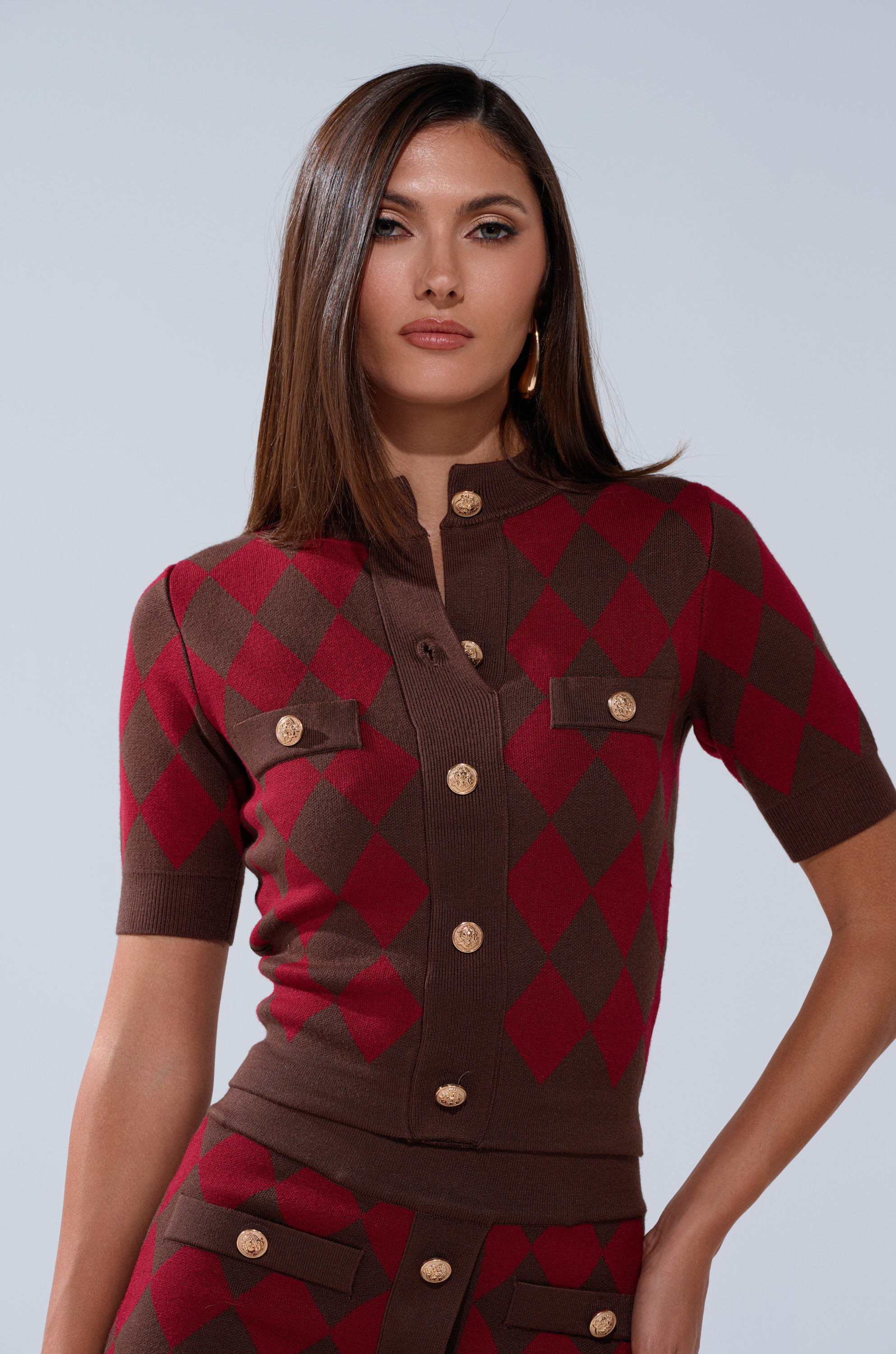 A woman with straight brown hair confidently models THE BEST PART OF THIS ABSTRACT CARDIGAN, a cropped knit design with gold buttons and a matching skirt, set against a plain light background.
