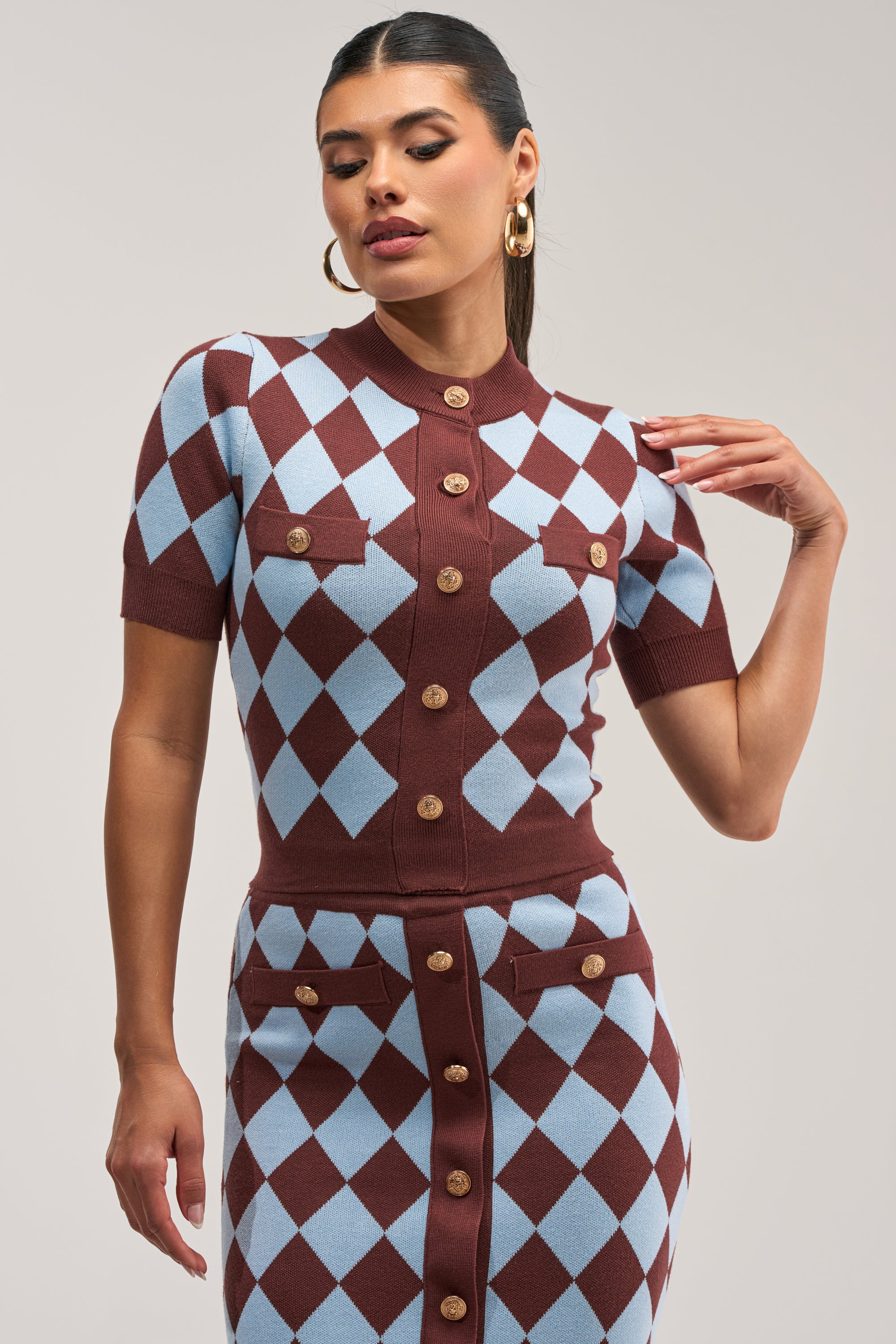 A woman poses against a plain background wearing the SWEET BABY GIRL BUTTON DOWN SWEATER, a short-sleeved dress featuring a blue and brown diamond pattern, gold buttons, and two chest pockets for a preppy-chic look.