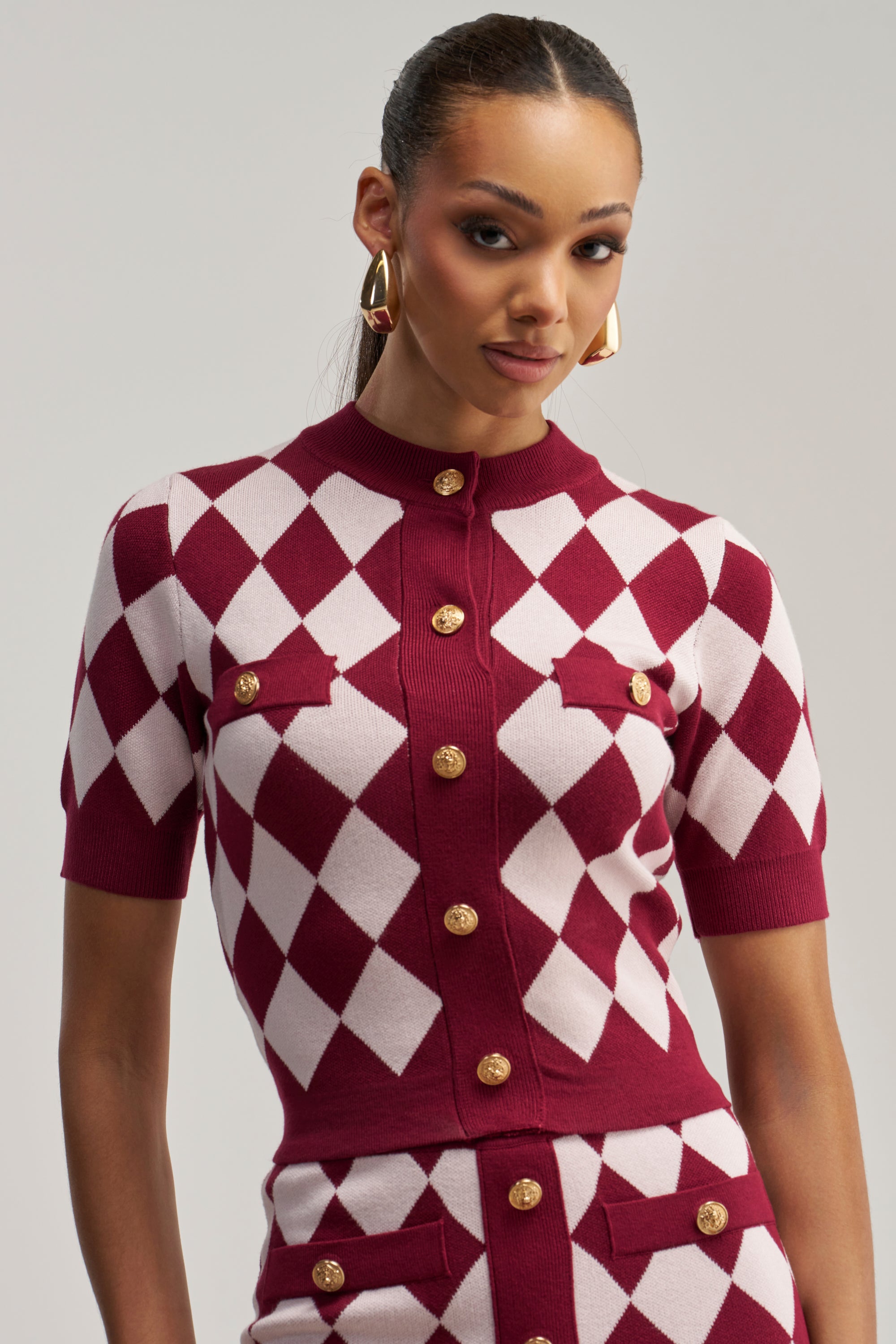 Dressed in the SWEET BABY GIRL BUTTON DOWN SWEATER IN RED MULTI, a woman models this cropped cardigan featuring a burgundy and white diamond pattern with gold button-down detail and a matching skirt. She completes her look with gold hoop earrings while posing against a neutral background, making this ensemble ideal for business casual occasions.