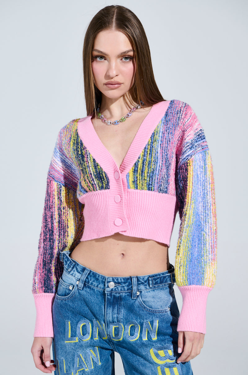 A woman showcases a COLOR POP CROPPED CARDIGAN featuring pink trim and buttons, paired with blue jeans with yellow text. Her long hair complements her necklace, while the cozy knit material stands out against a neutral backdrop.