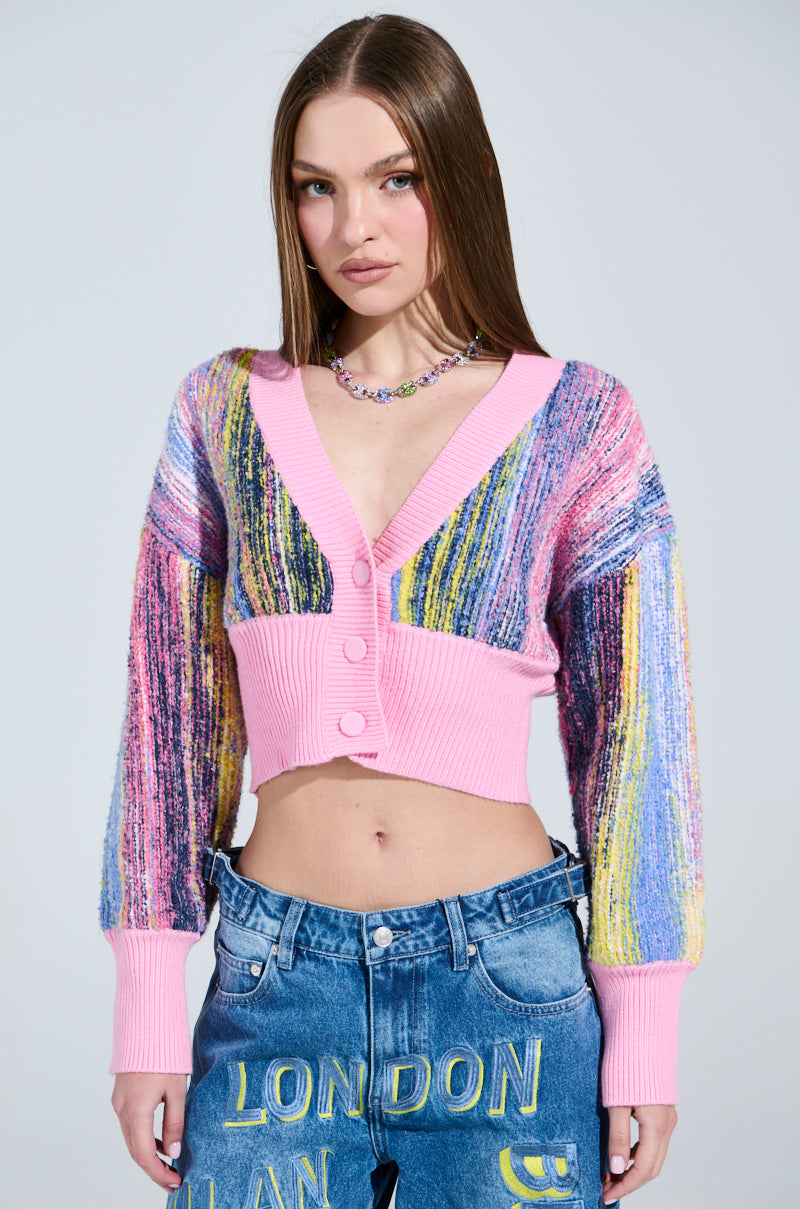 A person with long hair stands against a plain background, wearing the Color Pop Cropped Cardigan featuring pink buttons and cuffs. They pair it with blue jeans displaying "London" in yellow and accessorize with a multicolored necklace, showing a neutral expression.