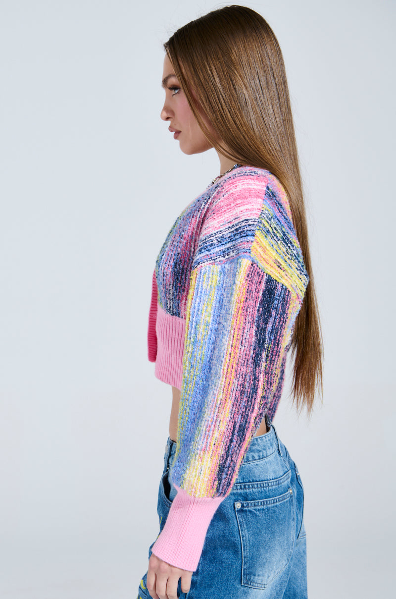 A person with long brown hair in side profile wears the COLOR POP CROPPED CARDIGAN—a cozy knit with pastel stripes of pink, blue, and purple. The long-sleeved sweater is paired elegantly with blue jeans against a plain light gray background.