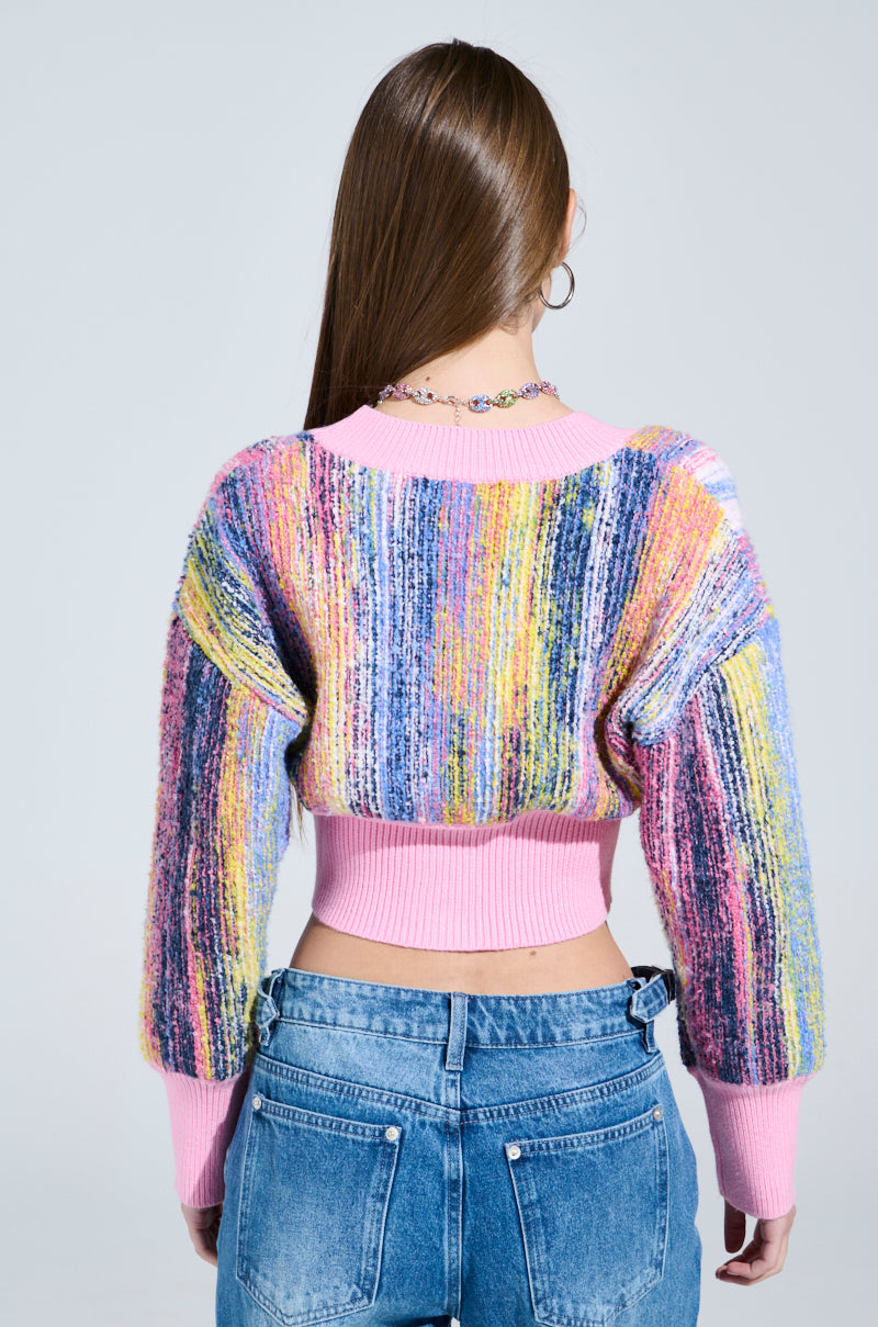 A person with long hair wears the COLOR POP CROPPED CARDIGAN, a pastel striped sweater with cozy knit material and pink cuffs and waistband, paired with light blue jeans, against a plain background.