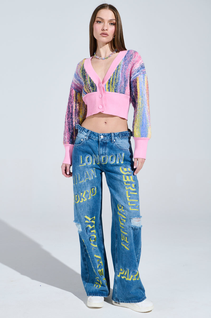 A person wears a cozy knit COLOR POP CROPPED CARDIGAN with multicolored sleeves and blue jeans featuring city names, set against a light gray background.