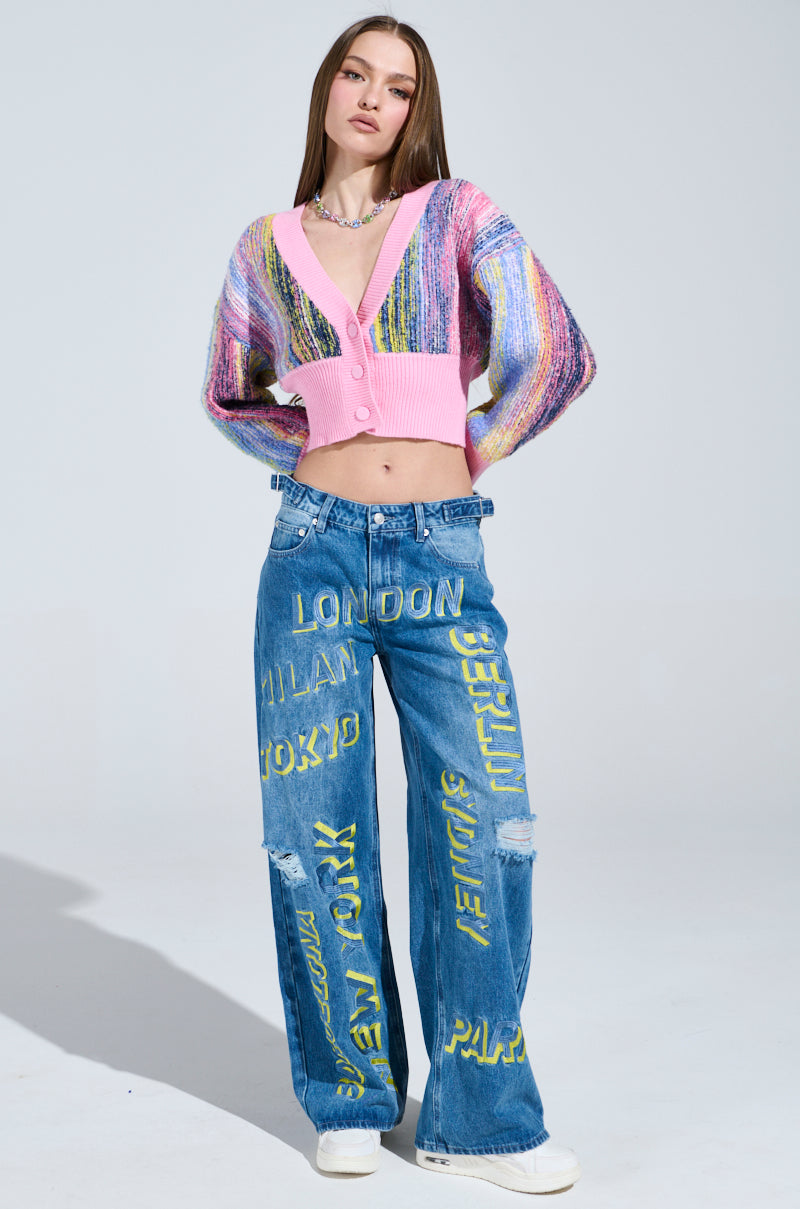 The individual confidently poses in a COLOR POP CROPPED CARDIGAN, featuring cozy knit and striped design. They're paired with blue jeans that display bold yellow city names and white sneakers, against a simple light gray backdrop.