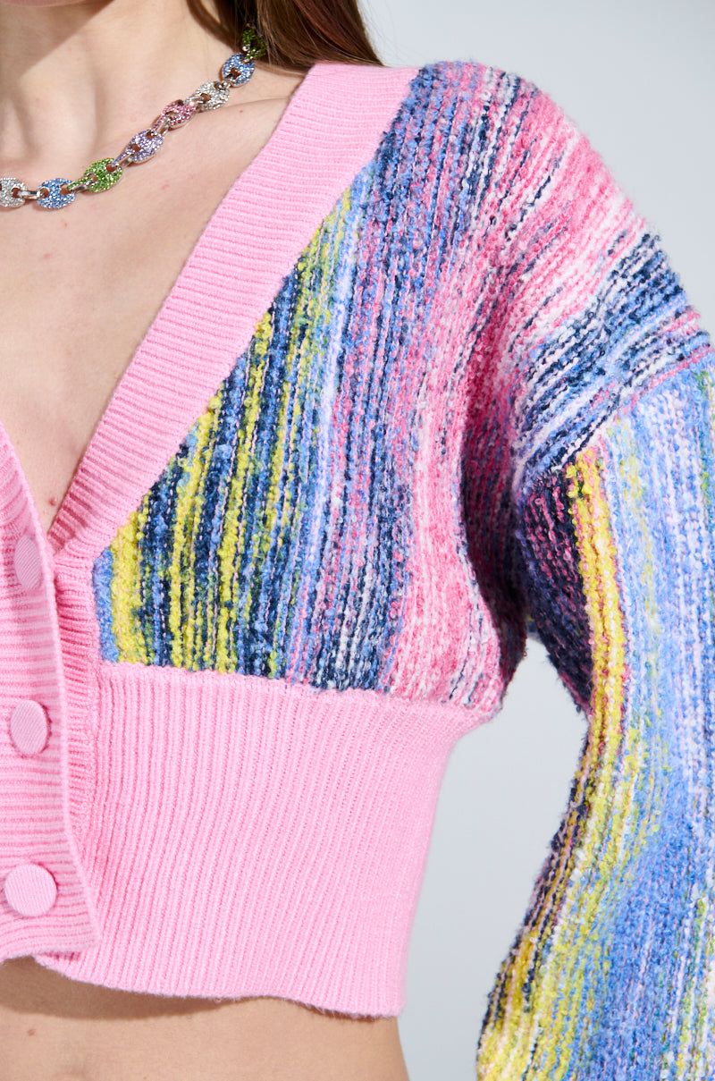 Close-up of someone in a COLOR POP CROPPED CARDIGAN, displaying pink, blue, yellow, and purple patterns. It features cozy knit material with a ribbed hem and large buttons. A floral necklace is partially visible around their neck.