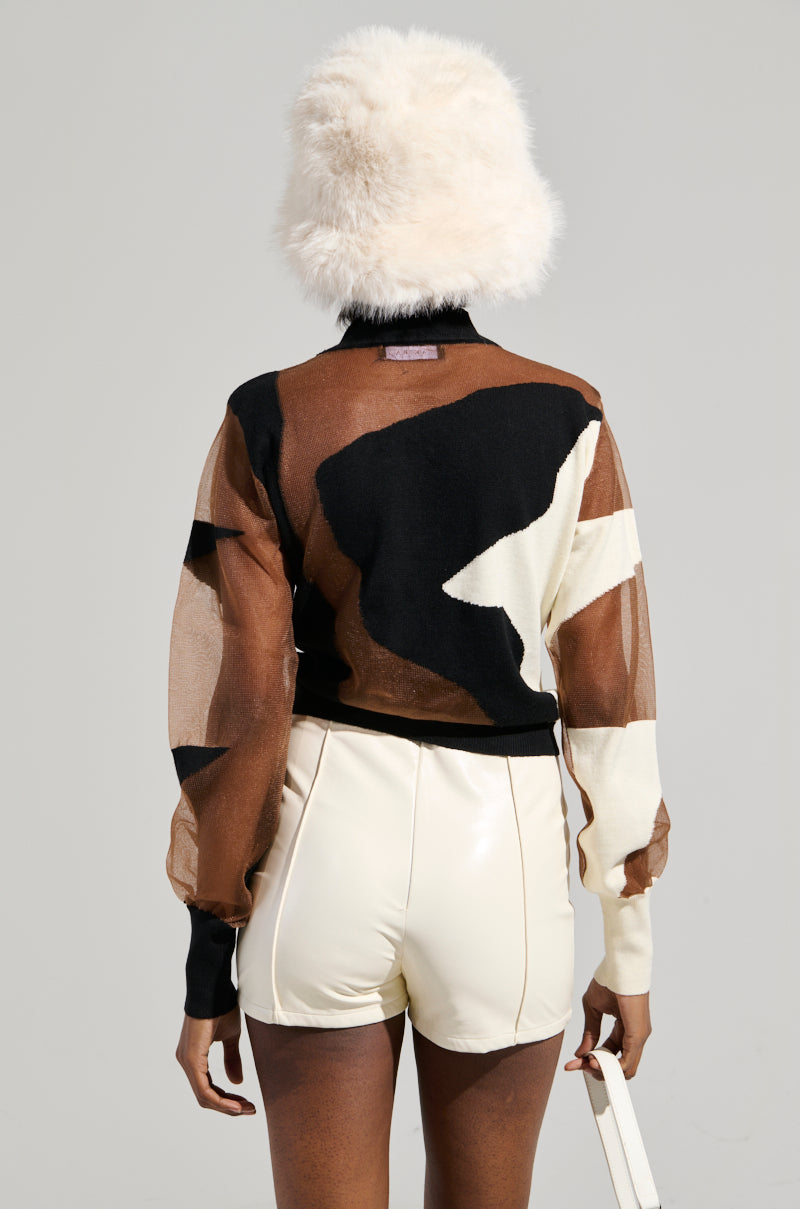 A person dressed in the BABY PLEASE MESH PANEL BLACK AND WHITE TOP featuring an abstract colorblock design and long sleeves, paired with light-colored high-waisted shorts and a fluffy white hat. The person is turned away from the camera against a plain backdrop.