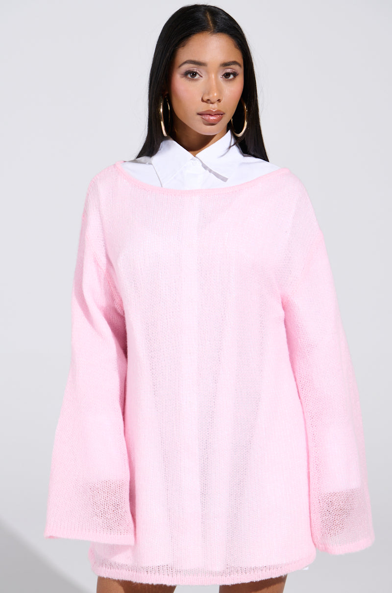 A person with long dark hair wears the BE READY IN 5 SWEATER DRESS in light pink, featuring a sheer, oversized fit over a white collared shirt. They stand against a plain background, looking forward with hands relaxed by their sides and wearing large hoop earrings.