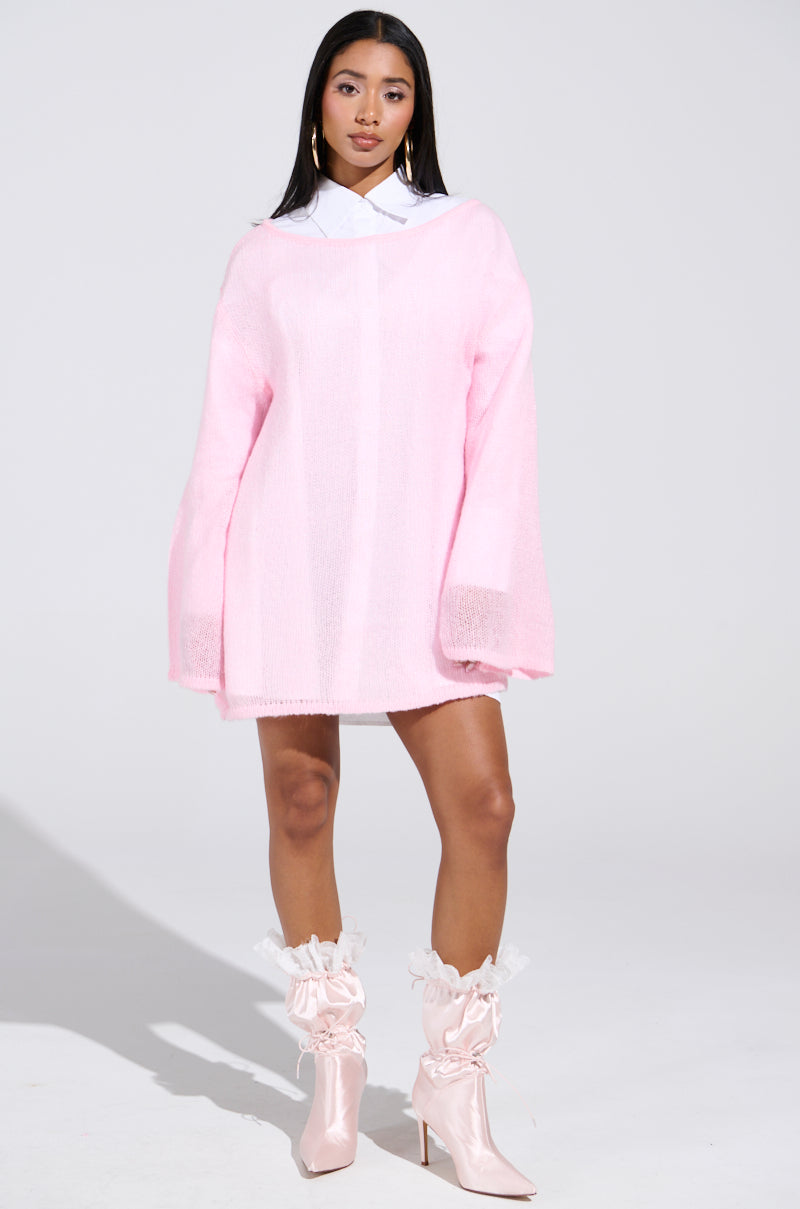 A woman stands against a plain background, wearing the BE READY IN 5 SWEATER DRESS—a light pink oversized sweater dress with a crisp white collar—and pairs it with light pink satin boots featuring ruffled tops. She gazes forward with a neutral expression.