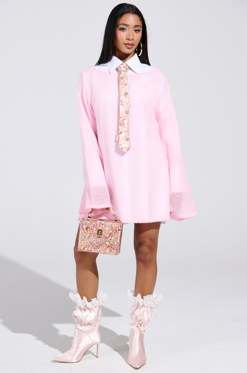 A person wears the BE READY IN 5 SWEATER DRESS, pink and oversized with bell sleeves, a wide white collar, and floral tie. They carry a sparkling handbag and wear shiny pink ankle boots with ruffled tops against a plain light background.