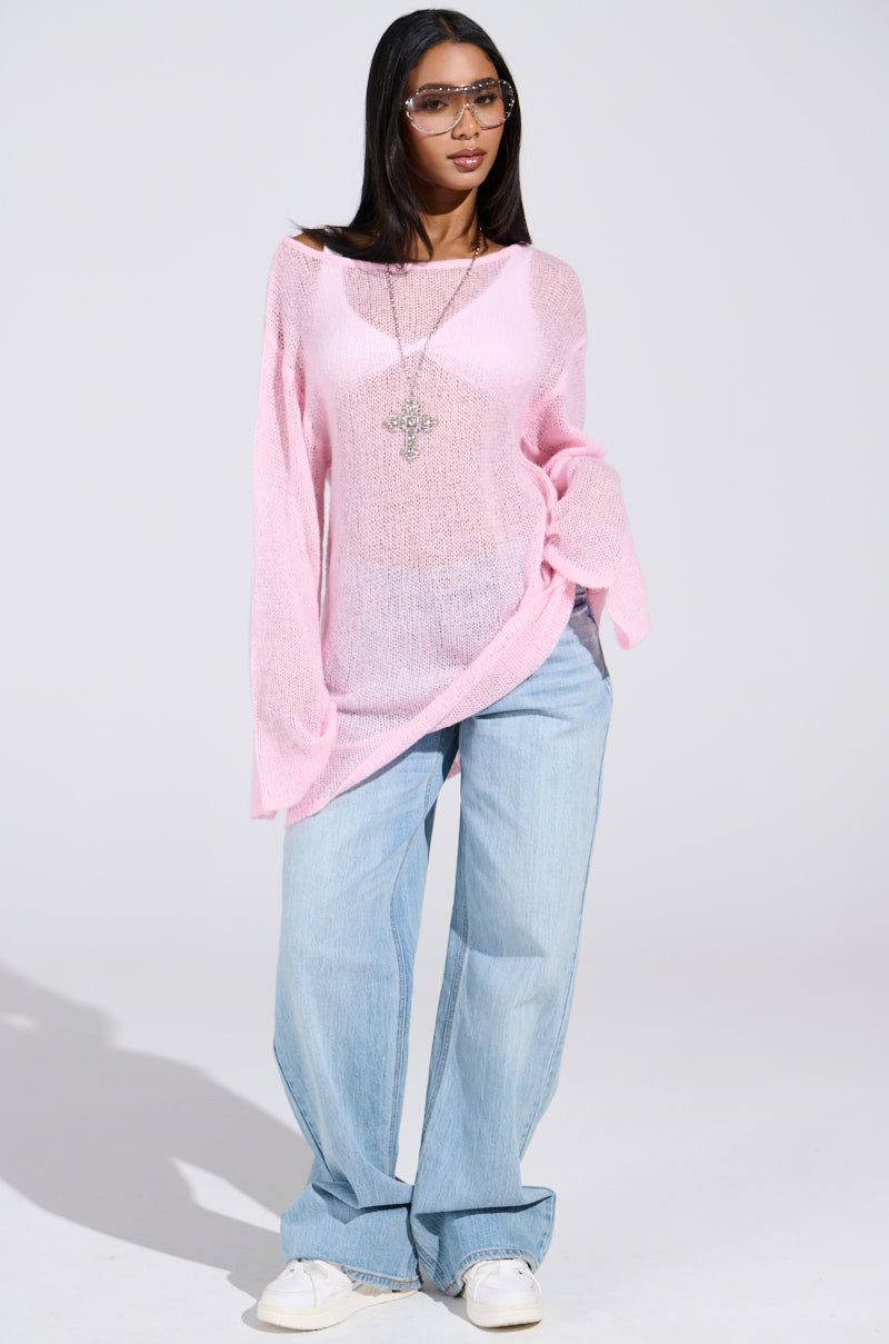 Confidently posing against a plain backdrop, a person wears the BE READY IN 5 SWEATER DRESS, an oversized sheer pink piece over a white top, paired with light blue wide-leg jeans and white sneakers. Sporting long hair, sunglasses, and a necklace complete their stylish look.