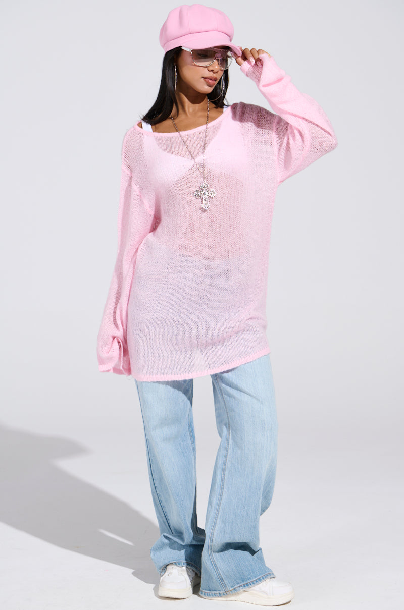 A person confidently wears a BE READY IN 5 SWEATER DRESS in sheer pink, paired with blue wide-legged jeans and white sneakers. They accessorize with a pink beret, sunglasses, and a long cross pendant necklace in front of a plain light gray backdrop.