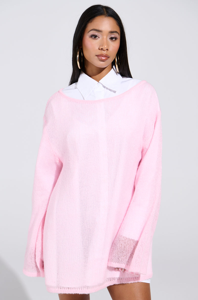 A person in the BE READY IN 5 SWEATER DRESS, light pink, over a white collared shirt stands against a neutral backdrop. They have long dark hair and wear large gold hoop earrings.