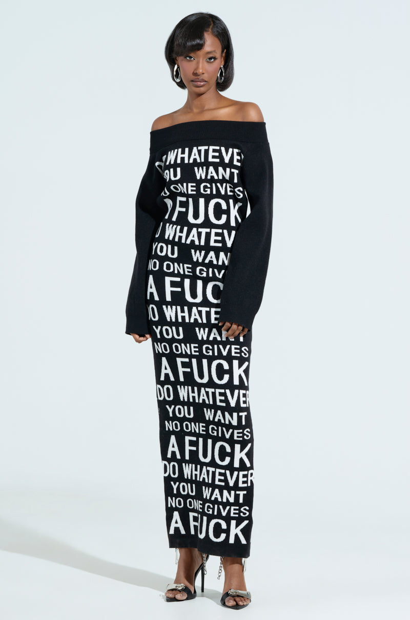 A person models the WHATEVER YOU WANT KNIT MAXI DRESS, a long off-the-shoulder black dress featuring repeating white text: "No one gives a fuck do whatever you want." They have short hair, wear hoop earrings, and high-heeled sandals against a plain white background.