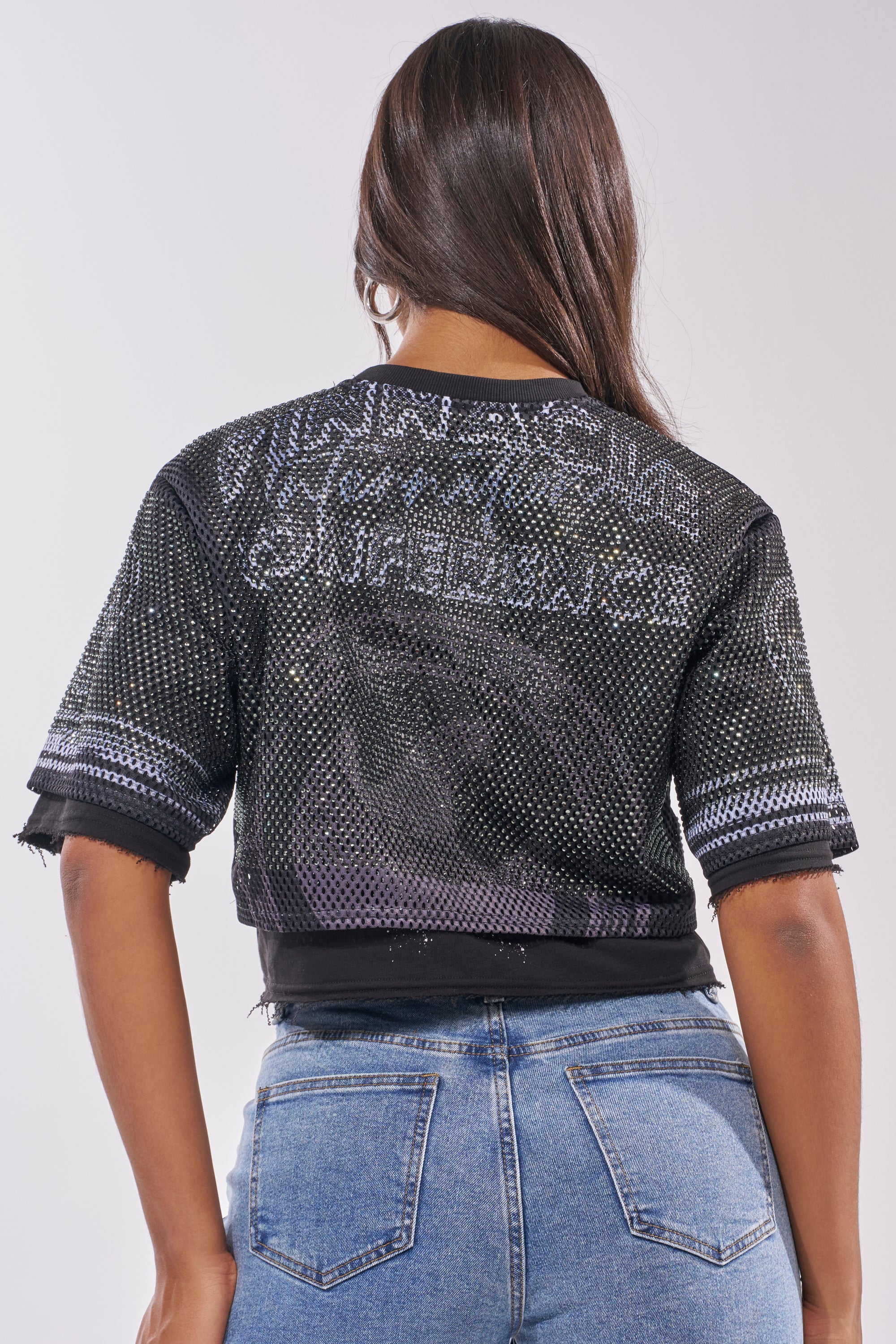 A woman with long brown hair wears the HIDDEN GEM RHINESTONE CROPPED TEE, a black sparkling mesh jersey with white writing, paired with blue jeans. She is seen from the back against a plain background. 3