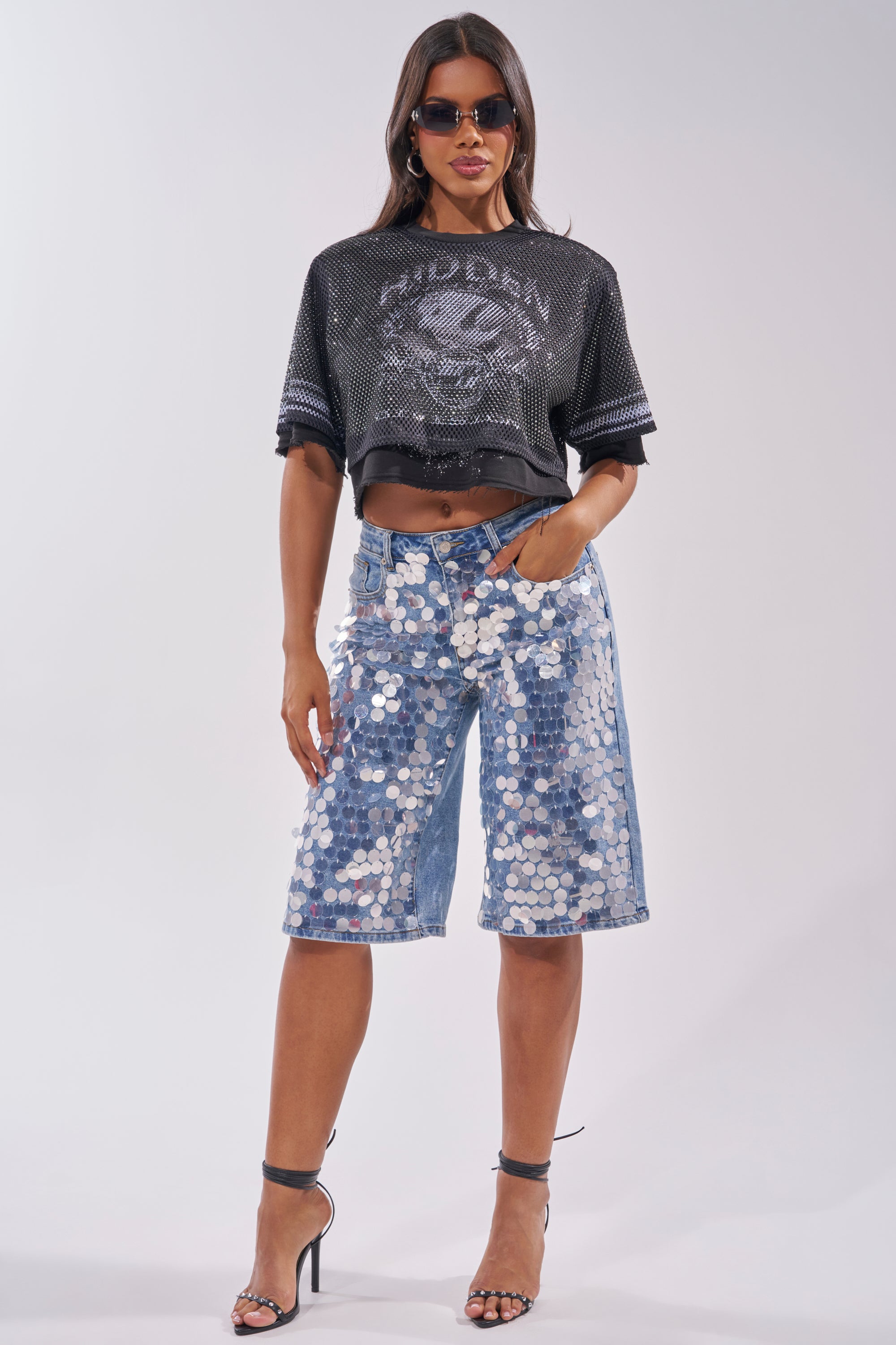 A woman wearing sunglasses, the HIDDEN GEM RHINESTONE CROPPED TEE, sequined knee-length denim shorts, and black strappy high heels stands confidently against a plain background. 4