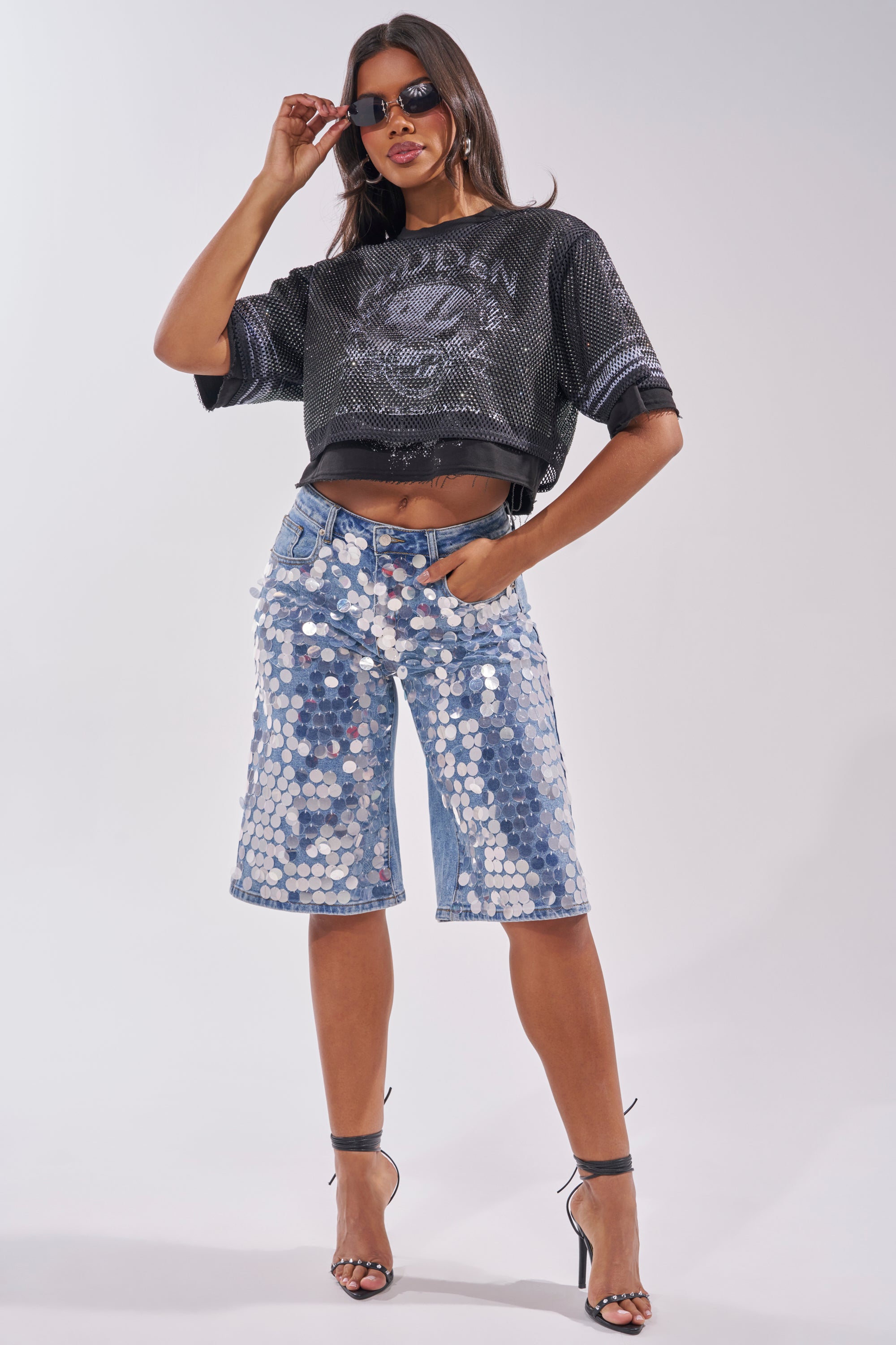 A woman in sunglasses and high heels poses confidently in the HIDDEN GEM RHINESTONE CROPPED TEE, a sparkly black mesh jersey, paired with knee-length denim shorts with large silver sequins against a plain background. 5