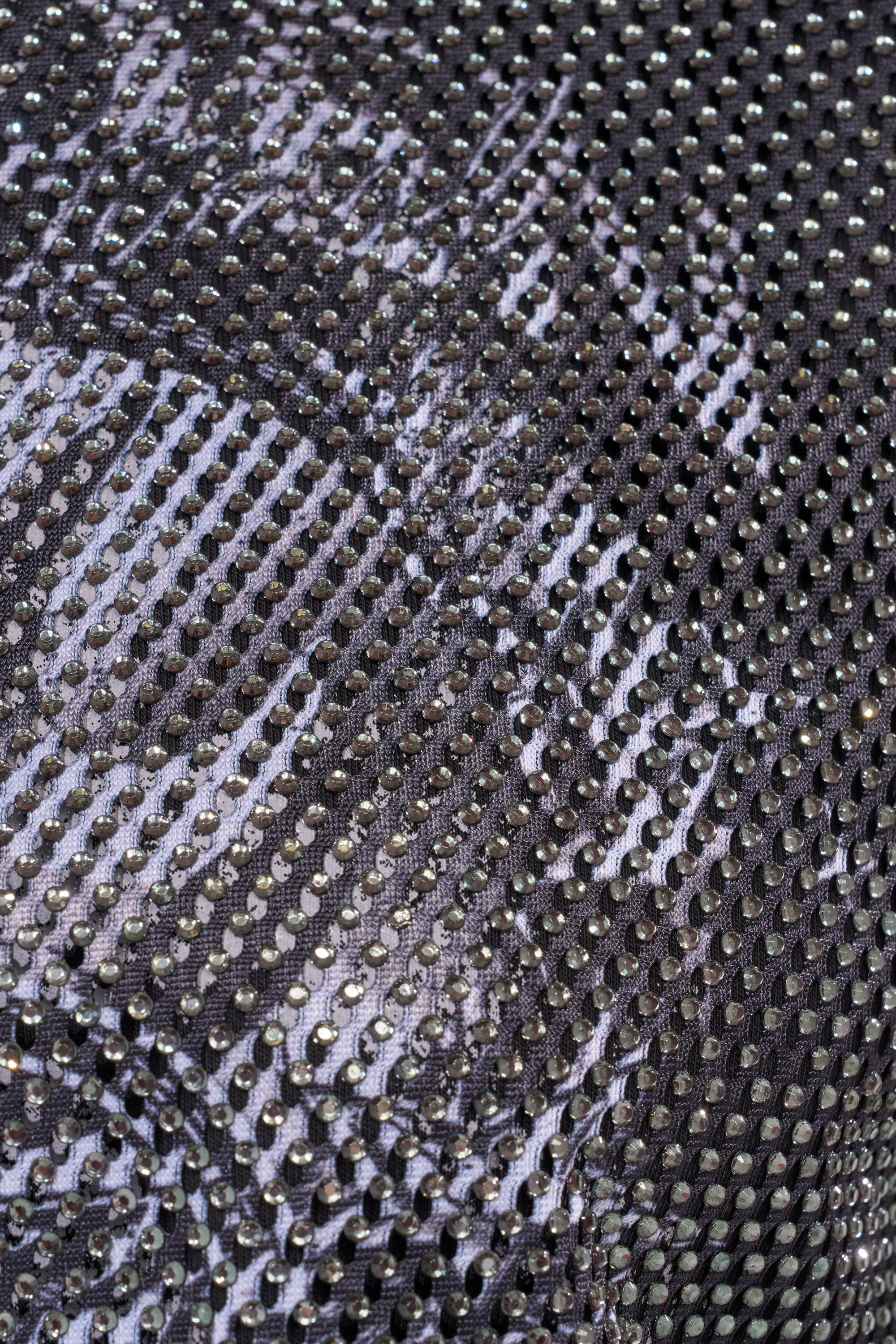 Close-up of the HIDDEN GEM RHINESTONE CROPPED TEE, showcasing shiny, reflective material with a black mesh overlay and small circular holes. The silvery metallic sheen gives it standout streetwear appeal. 6