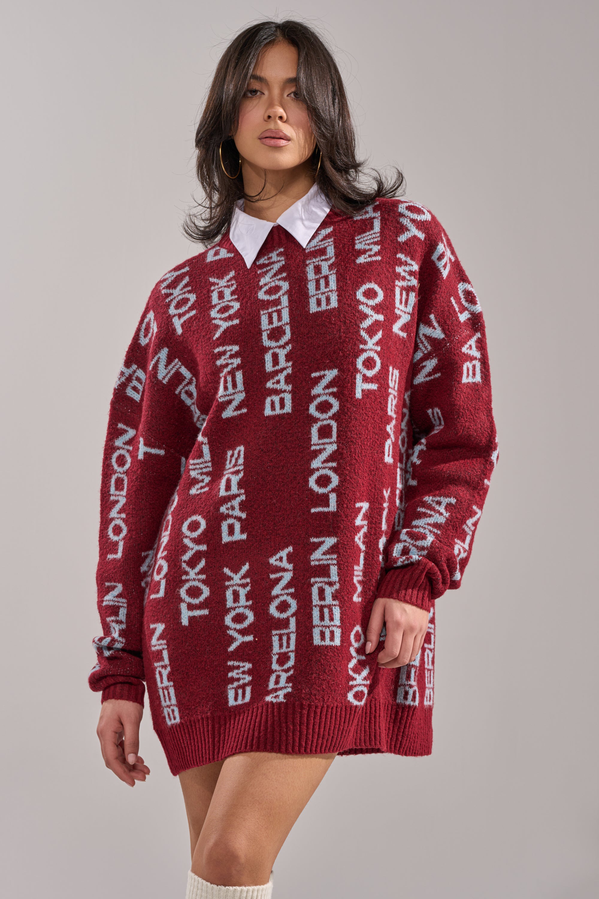 She wears the AROUND THE WORLD SWEATER MINI DRESS IN RED, featuring white city names all over. Styled with a white collared shirt underneath, wavy hair, hoop earrings, and a classic crew neckline. 0