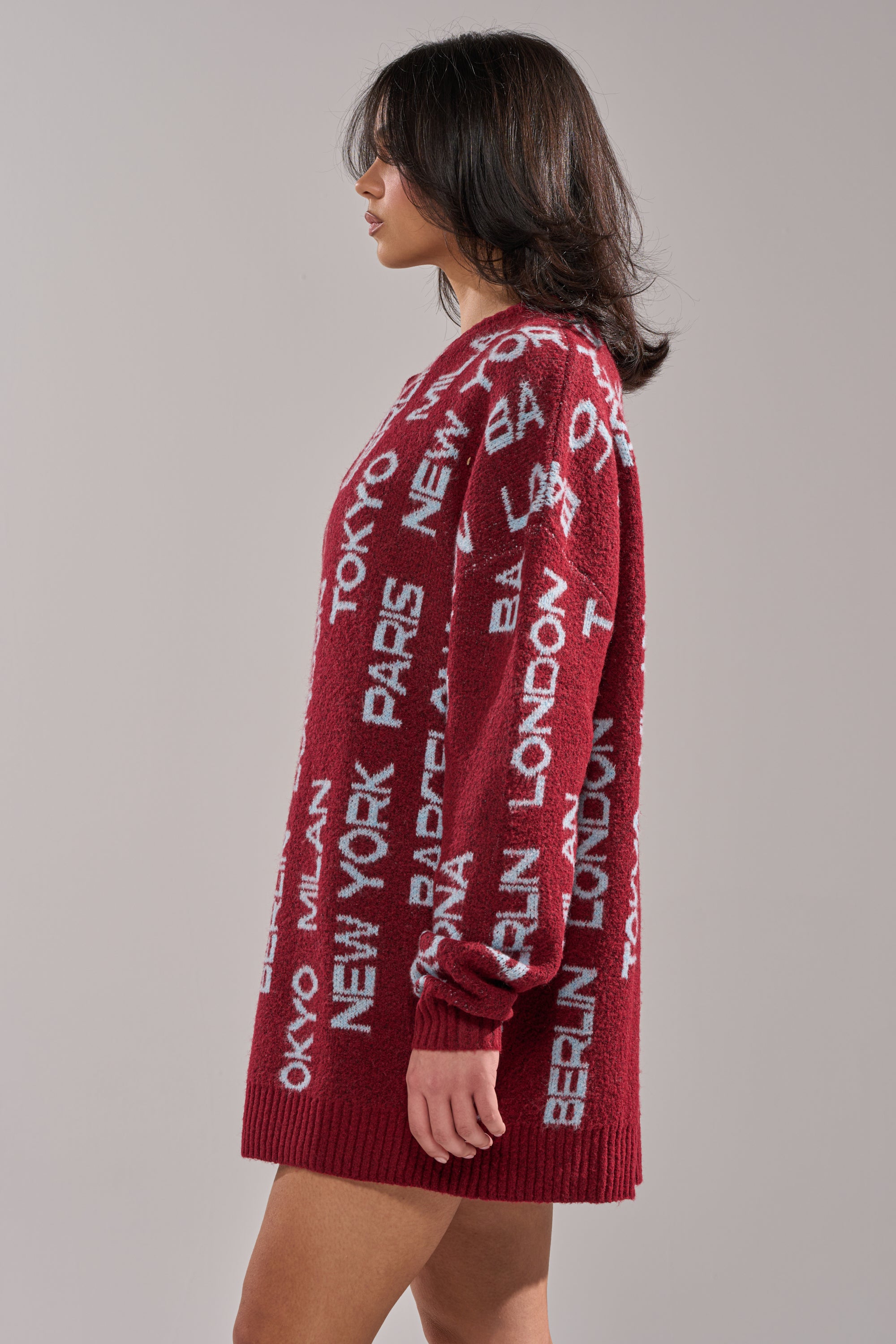 A woman with shoulder-length dark hair wears the AROUND THE WORLD SWEATER MINI DRESS IN RED, a long-sleeved knit featuring white city names. Seen in profile against a plain background, her look combines winter comfort and style. 2