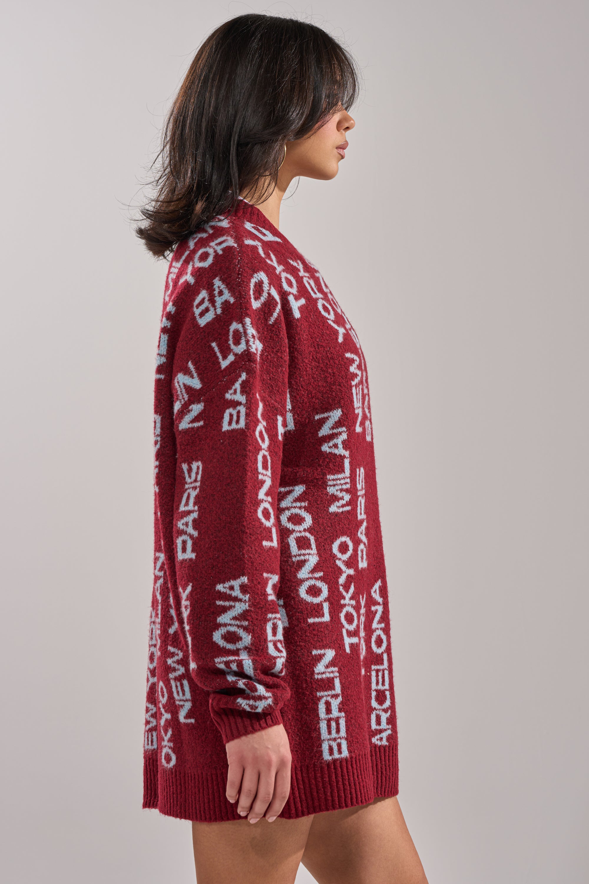 A woman with shoulder-length dark hair wears the AROUND THE WORLD SWEATER MINI DRESS IN RED, featuring white city name text, as she stands in profile against a plain light gray background. 3