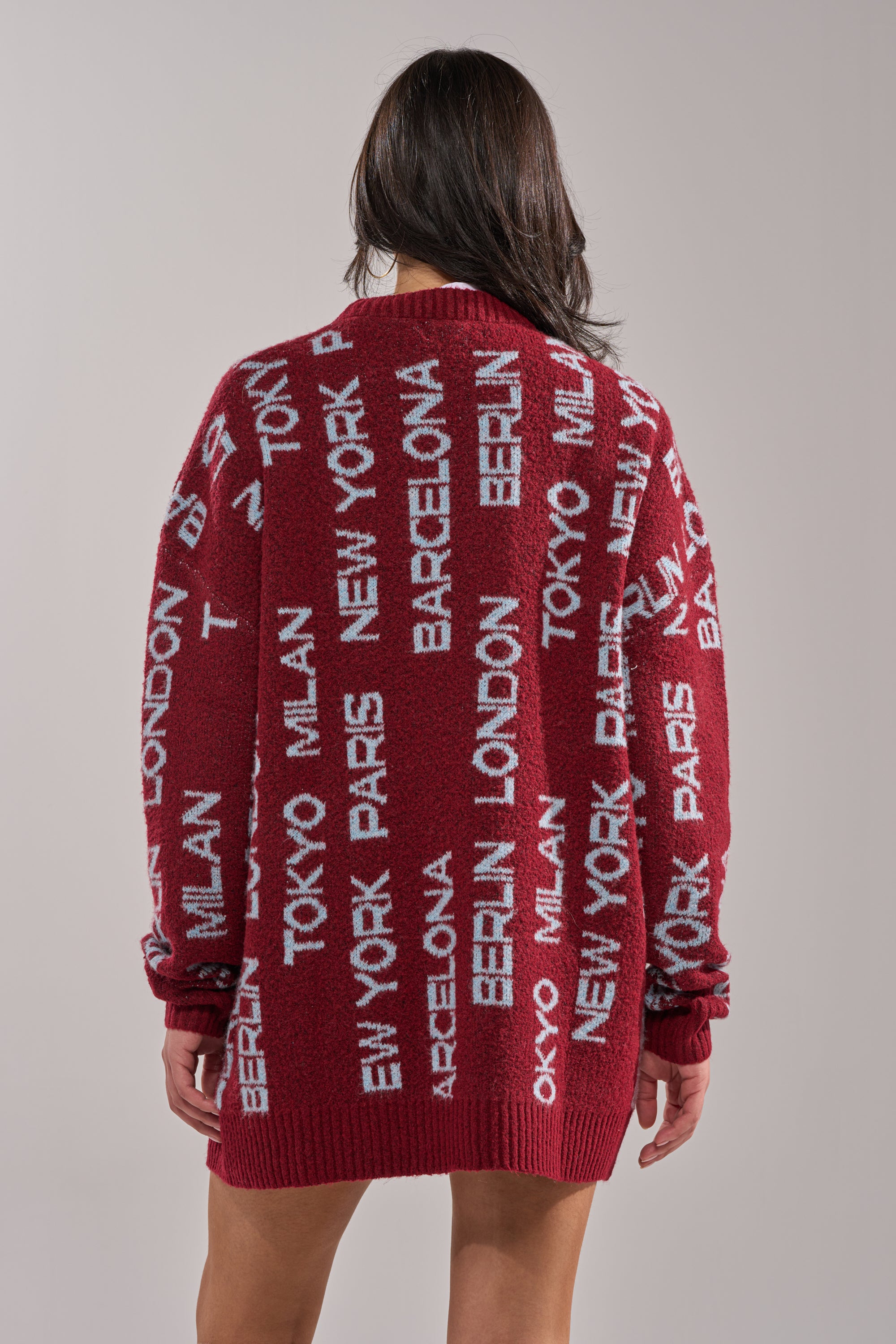 A woman with long brown hair faces away, wearing the AROUND THE WORLD SWEATER MINI DRESS IN RED, a loose winter pullover displaying major city names like New York, Tokyo, Paris, London, Milan, Barcelona, and Berlin in white block letters. 4