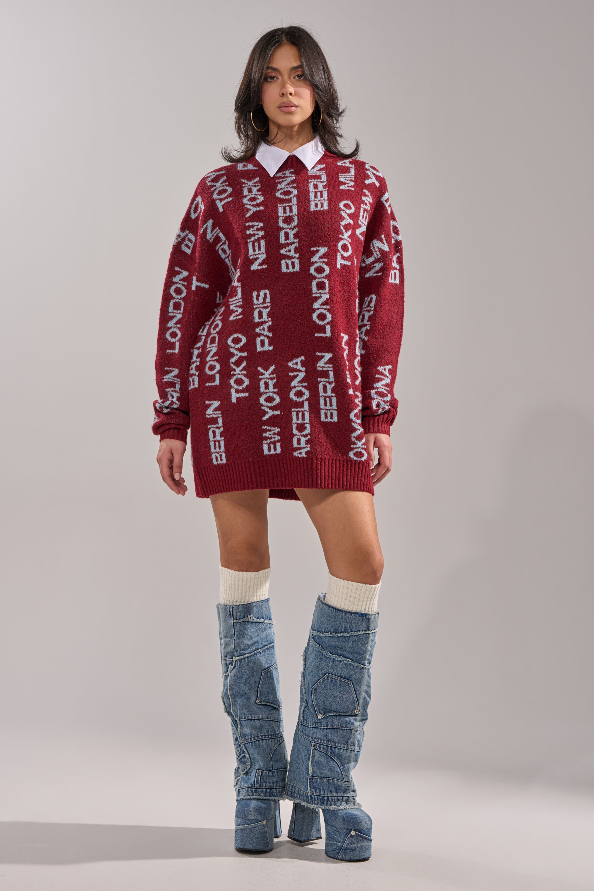 A woman wears the AROUND THE WORLD SWEATER MINI DRESS IN RED, featuring white city names, layered over a white collared shirt with tall slouchy denim platform boots. She poses against a plain light gray background. 5