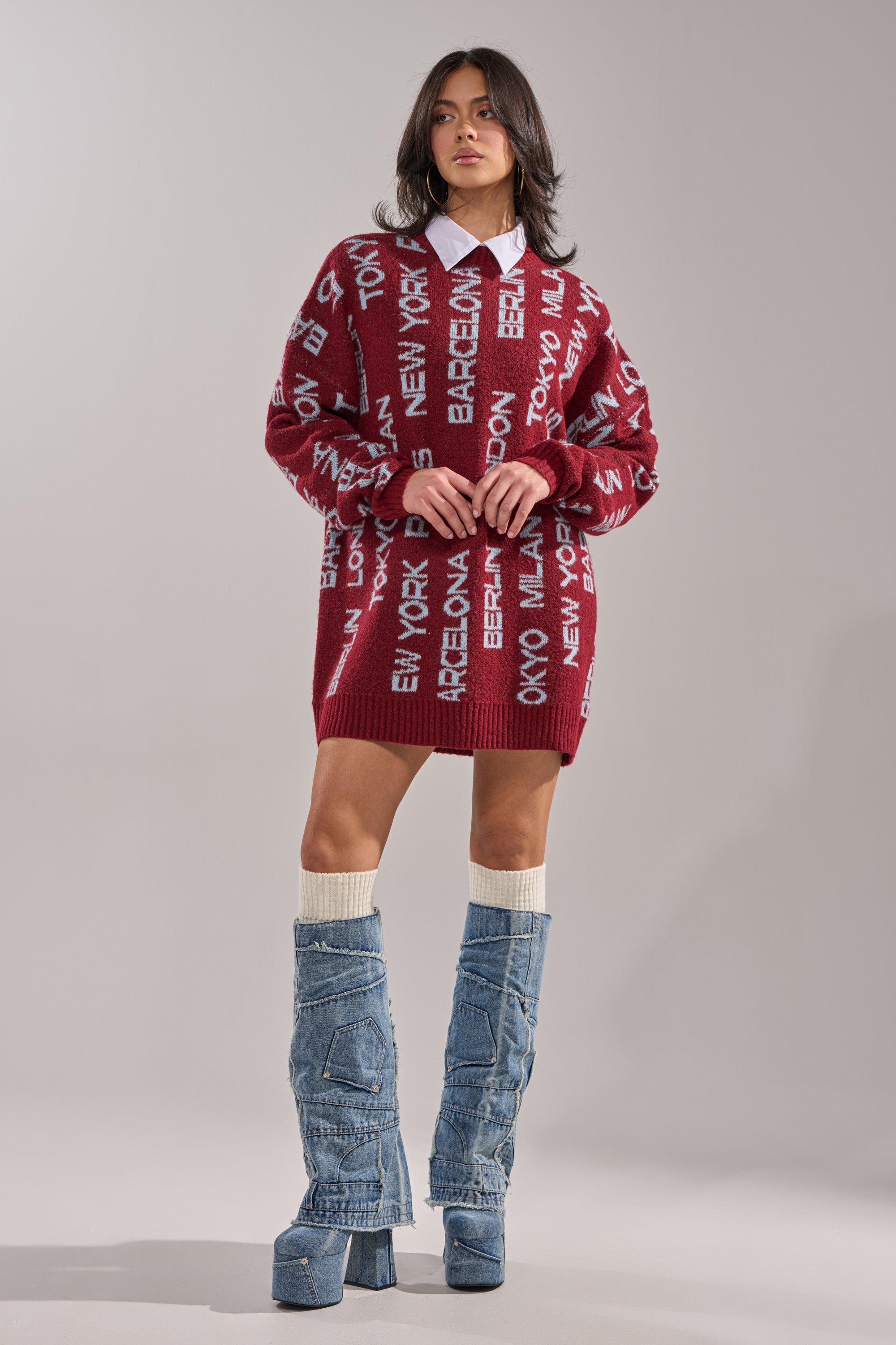 Wearing the AROUND THE WORLD SWEATER MINI DRESS IN RED over a white collared shirt and paired with light blue knee-high denim platform boots, a woman stands against a plain gray background. 6