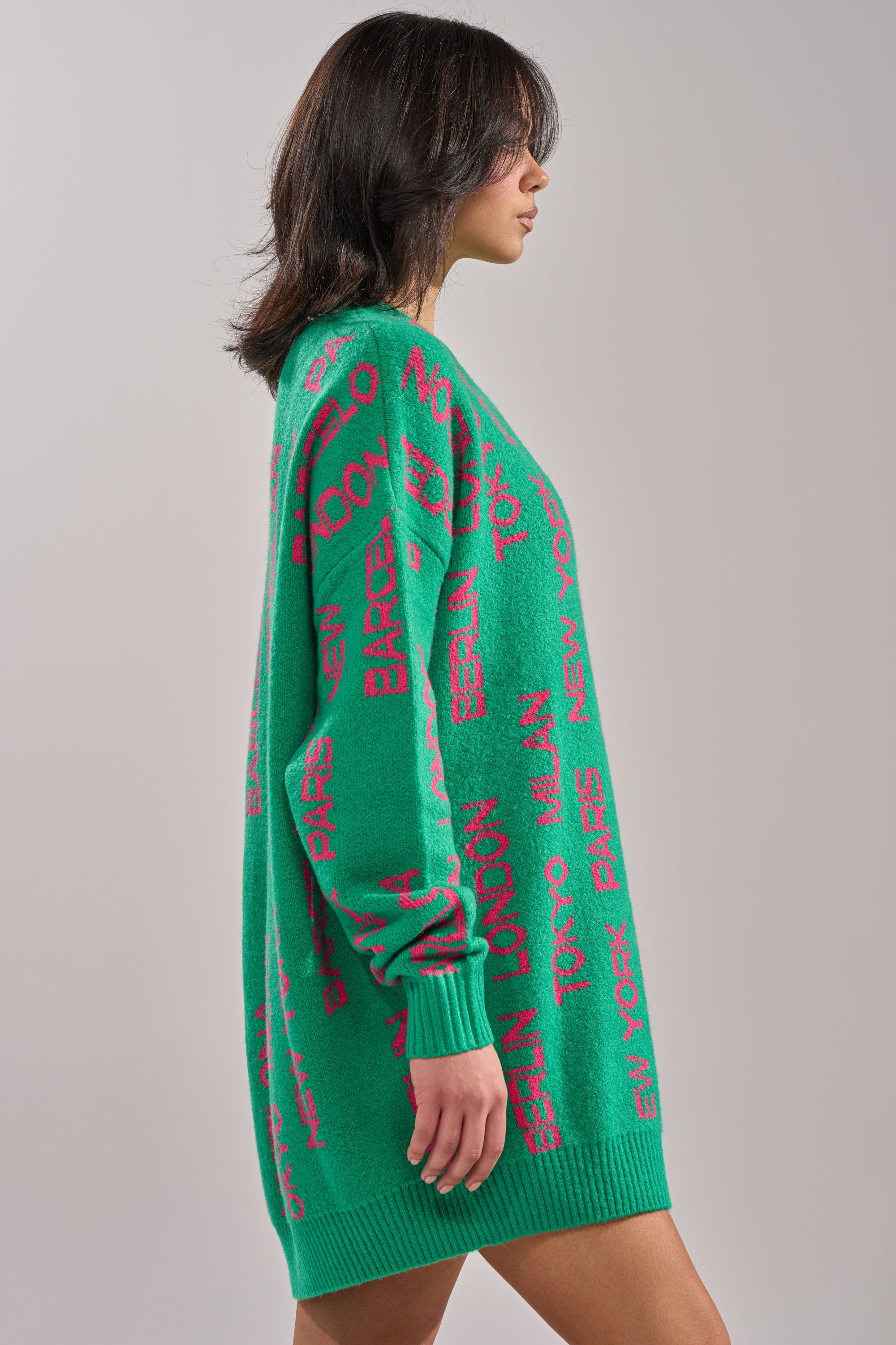 A woman stands sideways against a plain light background in the AROUND THE WORLD SWEATER MINI DRESS IN GREEN, featuring an oversized fit and red city names like New York, Tokyo, and Paris printed all over. 2