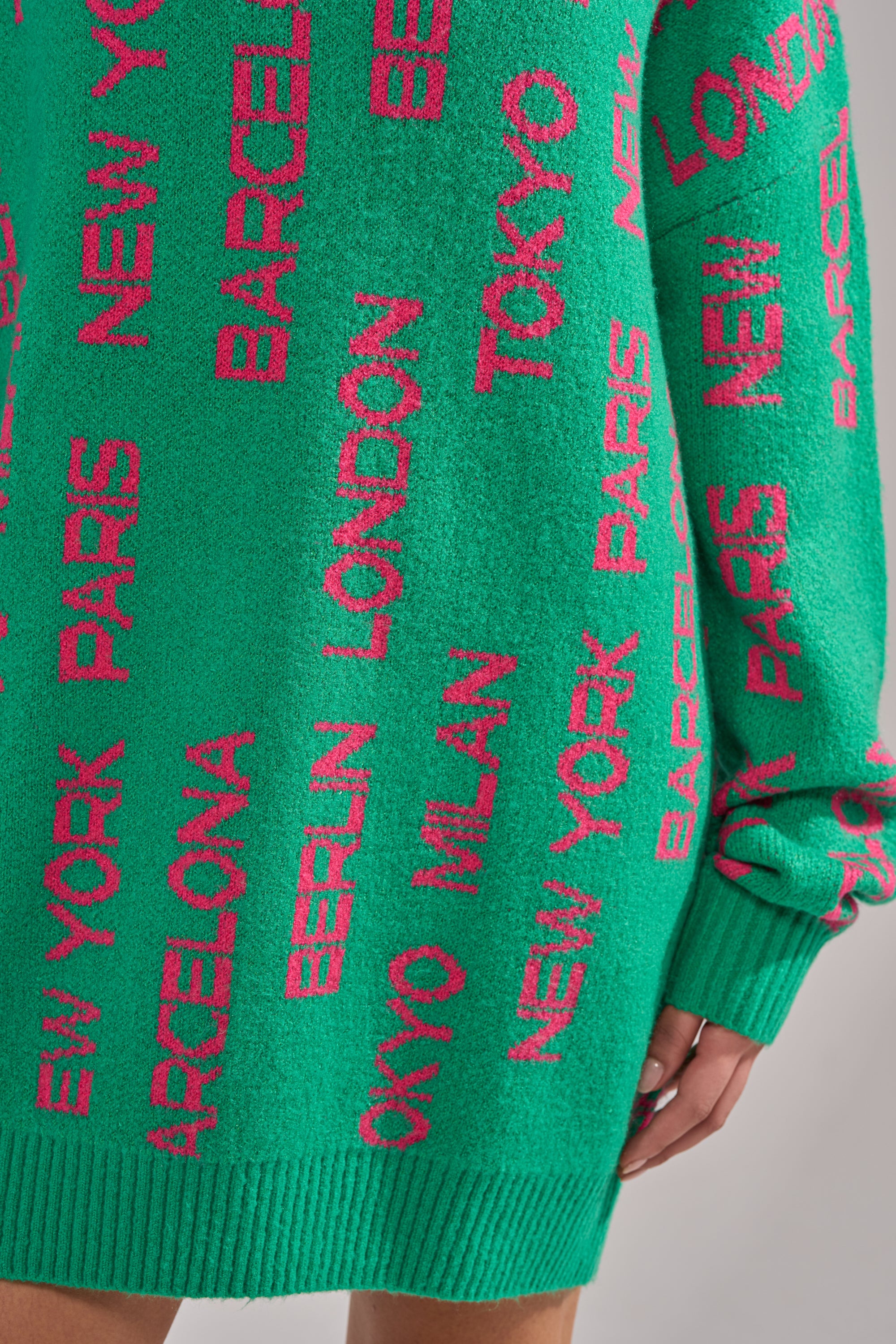 The AROUND THE WORLD SWEATER MINI DRESS IN GREEN features pink city names like New York, Paris, and Tokyo in various orientations on cozy knit fabric. This oversized mini dress is shown with part of a hand and leg visible. 6