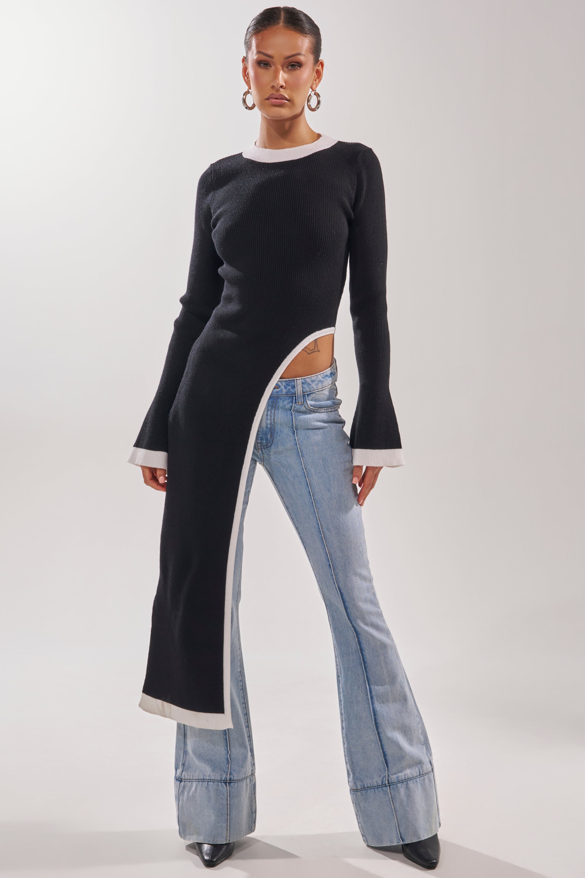 A woman models the SUPER SOFT EXTENDED SWEATER, a black asymmetrical piece with a white-trimmed hem, long on one side past her knee and short on the other to reveal her hip, paired with light blue flared jeans.