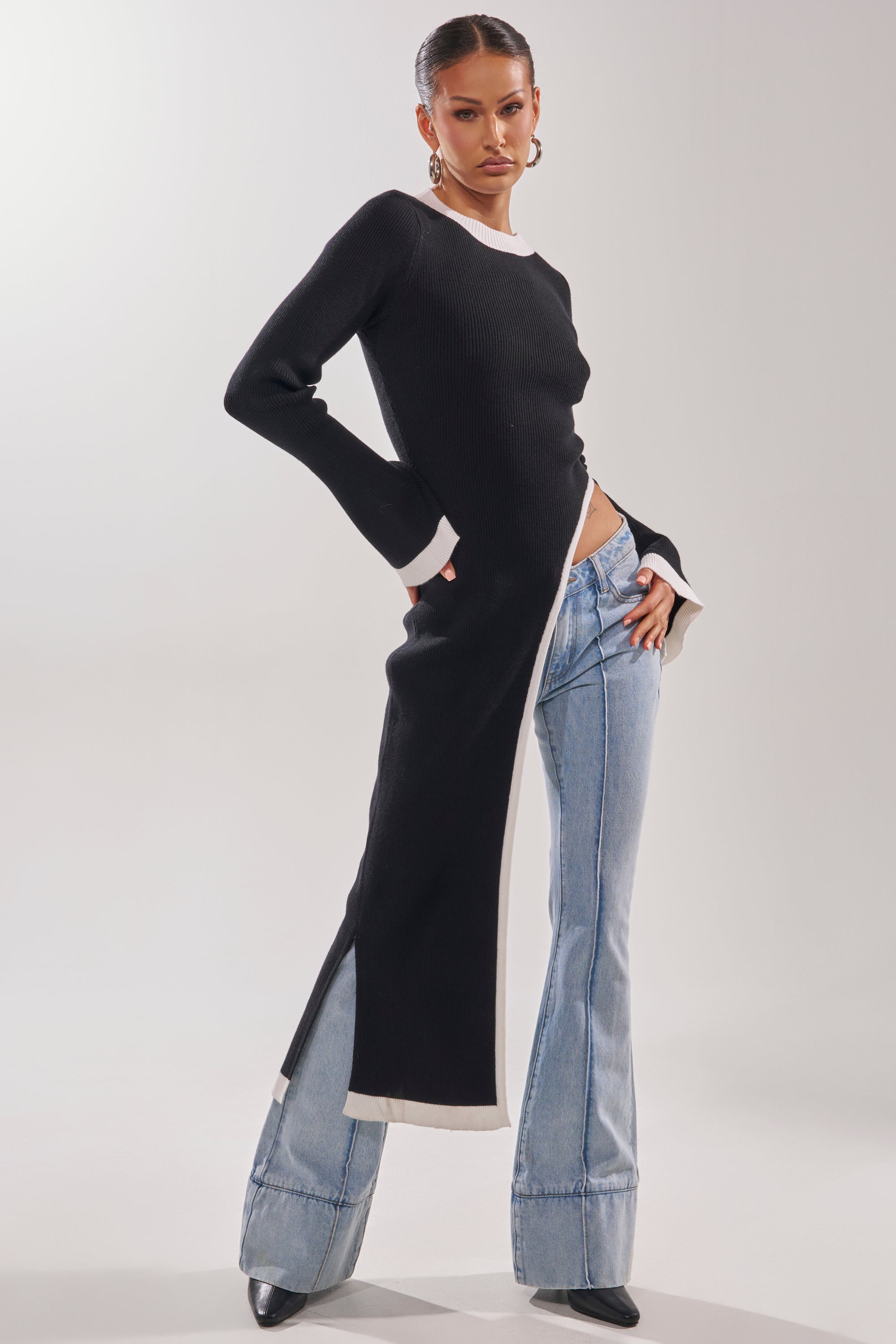 A woman poses confidently in the SUPER SOFT EXTENDED SWEATER, a black and white asymmetrical piece with high side slits, paired with light-wash flared jeans and black pointed-toe shoes against a plain background.