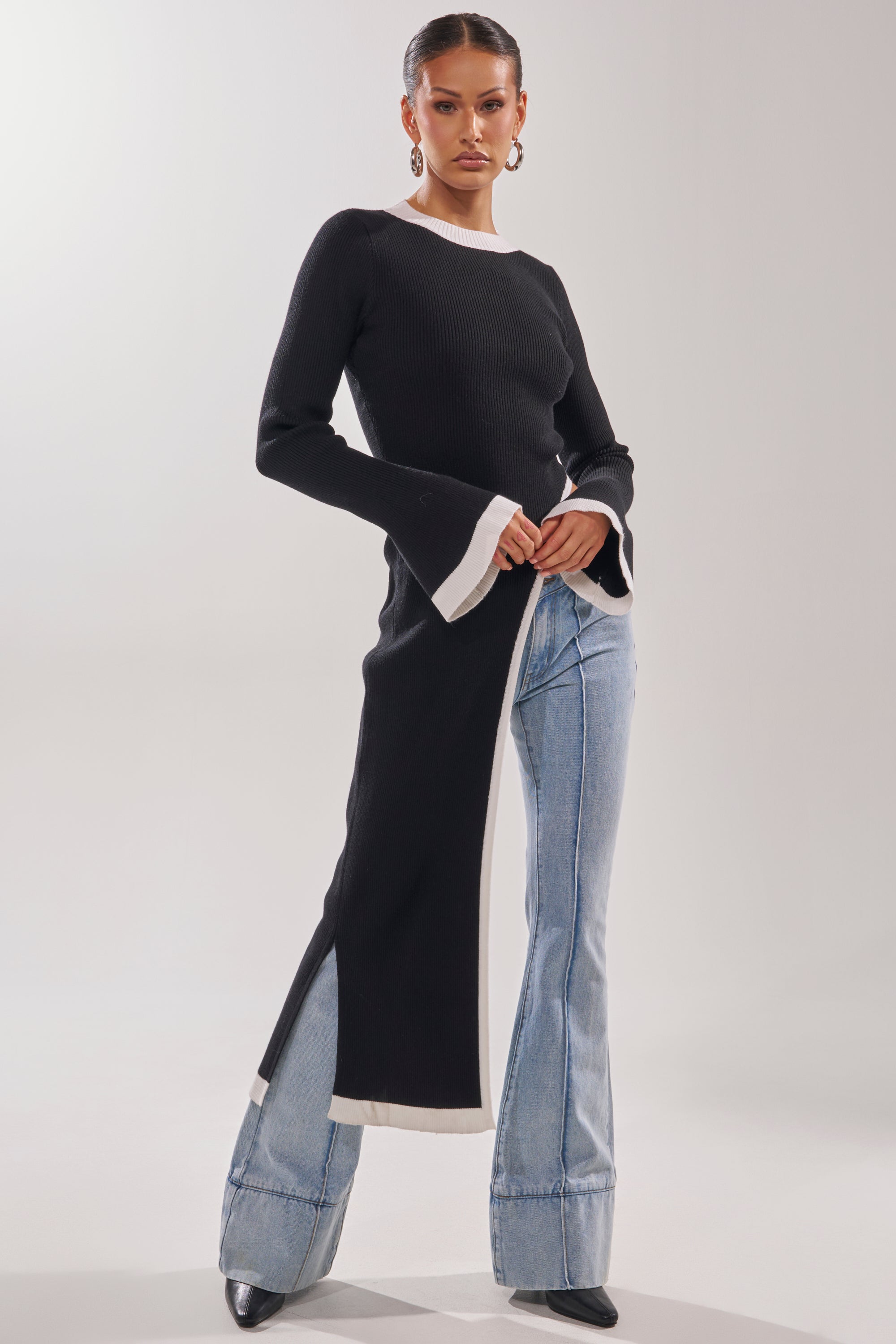 A woman stands confidently in the SUPER SOFT EXTENDED SWEATER with bell sleeves, paired with light blue flared jeans and black pointed-toe shoes. Her hair is slicked back and she completes the look with statement earrings.
