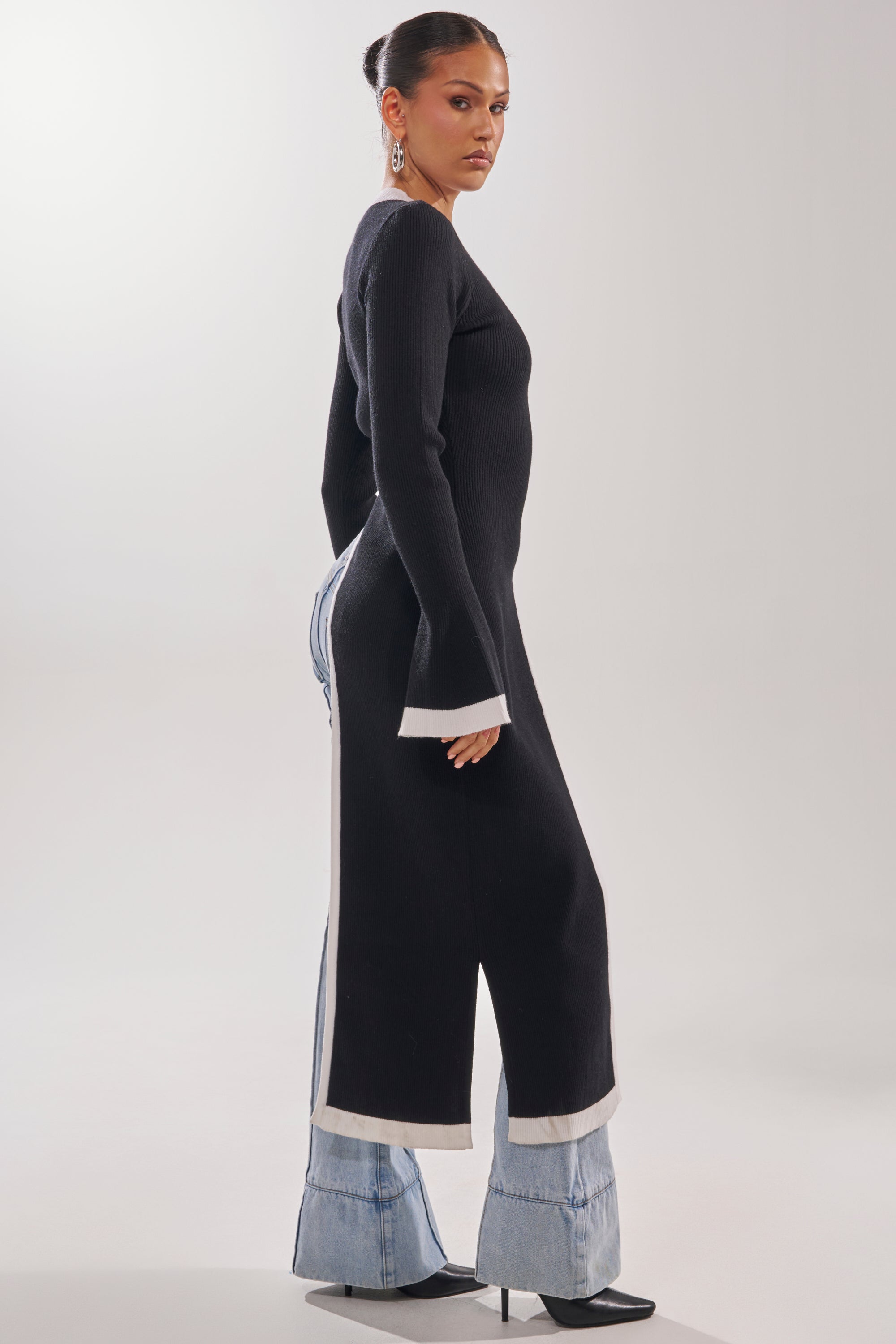 A woman poses sideways against a white background wearing the SUPER SOFT EXTENDED SWEATER—a long, black rib-knit dress with white cuffs—styled over wide-legged light-wash jeans and black pointed-toe heels.