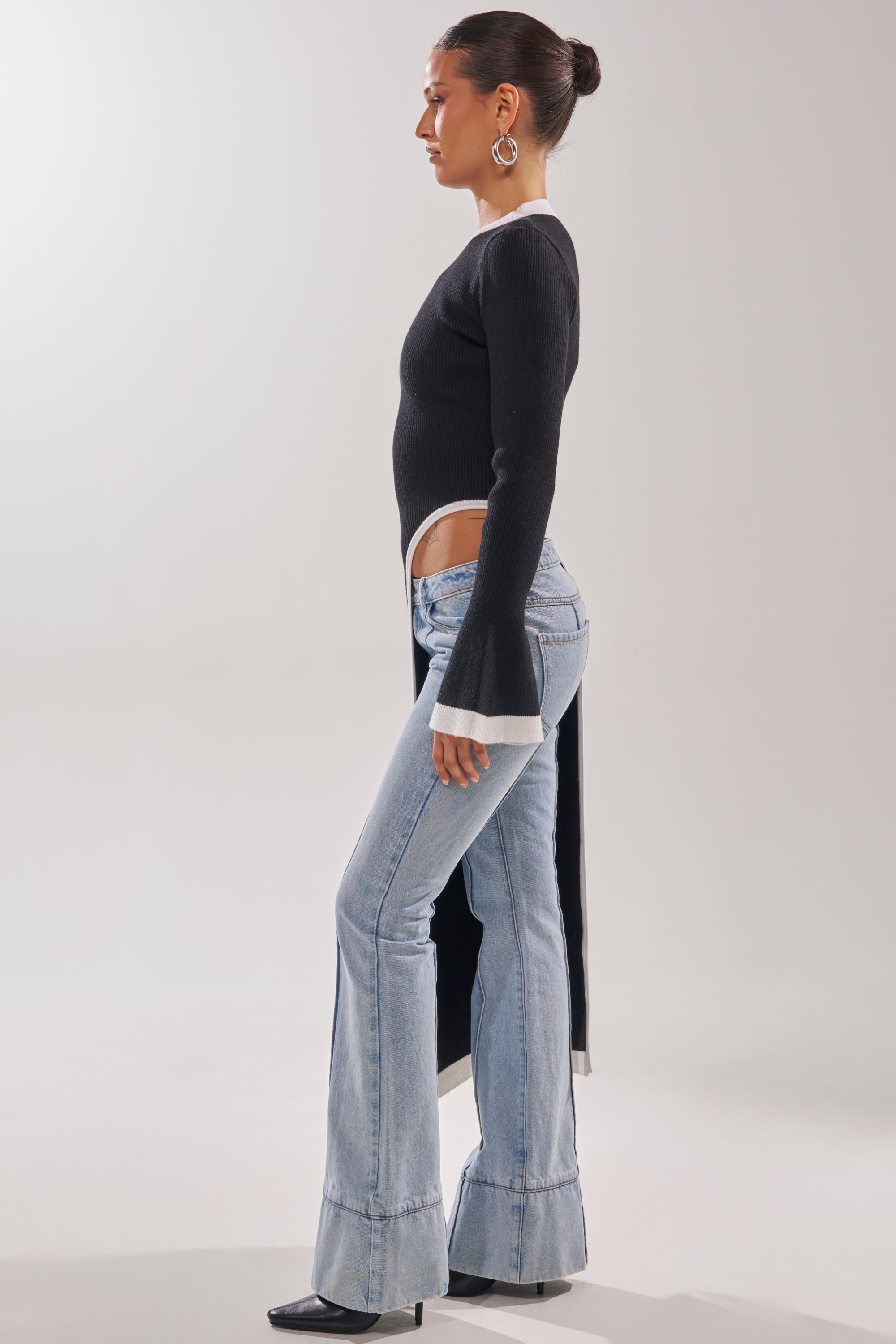 A woman in profile models the SUPER SOFT EXTENDED SWEATER with flared cuffs and a high slit, paired with light blue flared jeans, black heels, large hoop earrings, and her hair styled in a neat bun.