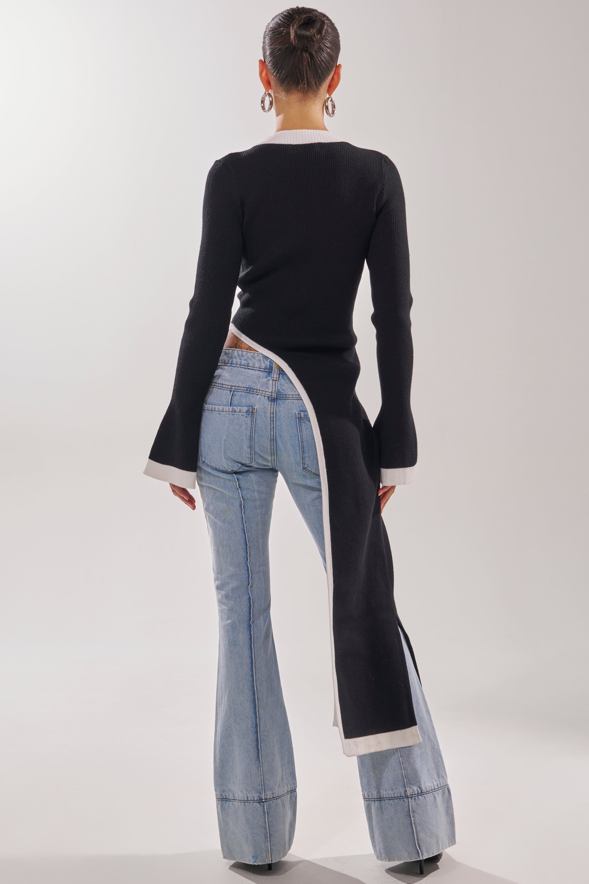 A person faces away in light blue flared jeans and the SUPER SOFT EXTENDED SWEATER—a black piece with white trim, an asymmetrical long split hem, paired with hoop earrings and a neat bun.