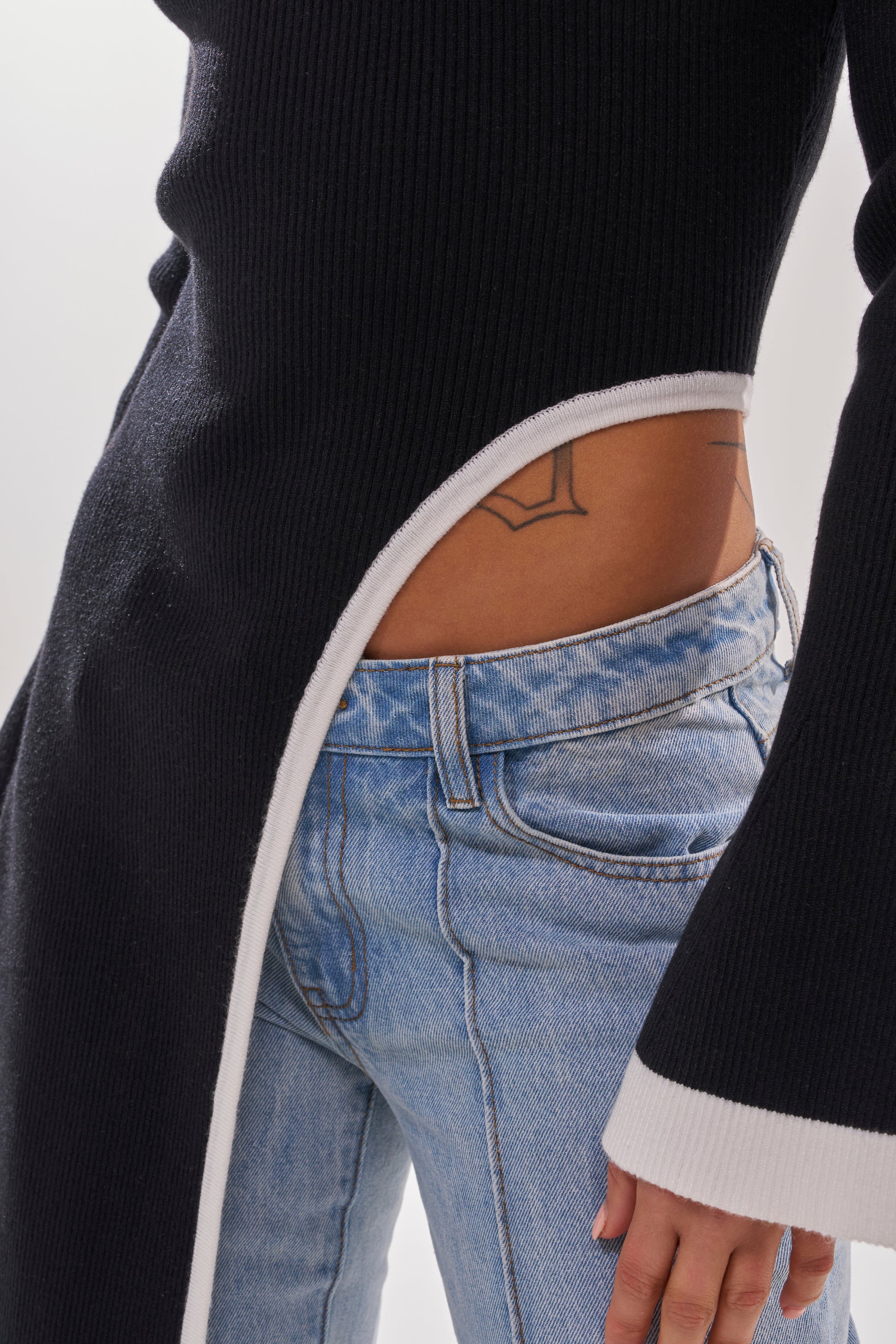 A person in light blue jeans and the SUPER SOFT EXTENDED SWEATER—a long-sleeve top with white trim, an asymmetrical midriff-baring cut, and a geometric waist tattoo visible.