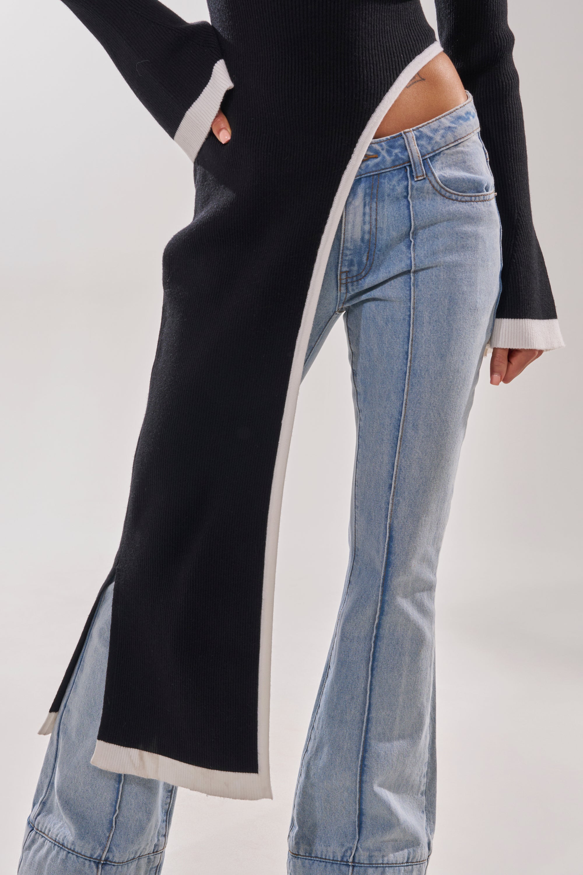 A person wears the SUPER SOFT EXTENDED SWEATER, featuring white trim, a high hip-revealing slit, and a sleek, super soft design paired with light blue flared jeans against a plain backdrop.