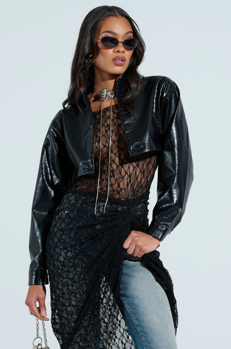 A woman poses confidently in sunglasses, sporting a chic ensemble with the NATALIA CROCODILE PU BUTTON DOWN SHIRT over a sheer lace top, paired with a long lace skirt layered over jeans. She holds a chain accessory, and her wavy hair complements the edgy look.