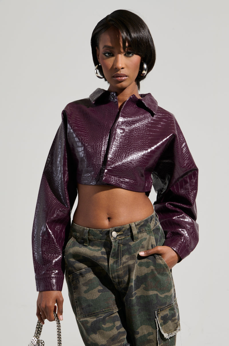Sporting a shiny Natalia Crocodile PU Button Down Shirt alongside camouflage pants, the person poses confidently. Their short hair and the chain strap they hold accentuate their bold fashion choice against the plain background.