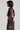 A person with a short bob haircut is wearing the NATALIA CROCODILE PU BUTTON DOWN SHIRT, which is a shiny, dark purple faux leather cropped jacket, paired with camo-patterned pants. The image captures the chic silhouette in profile against a plain background.