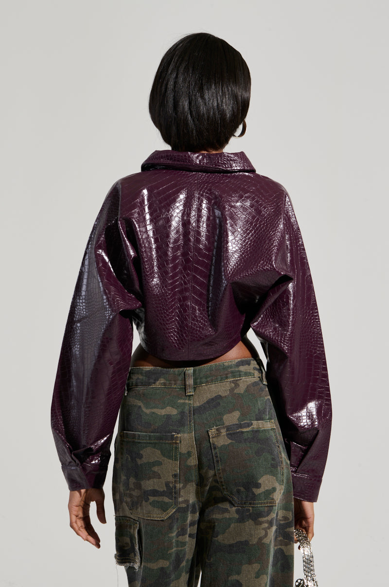 Rear view of a person wearing the NATALIA CROCODILE PU BUTTON DOWN SHIRT, featuring a dark purple, shiny textured finish with a cropped silhouette, paired with camouflage pants. The high-collared shirt with wide sleeves complements the person's short hair against the plain, light-colored background.